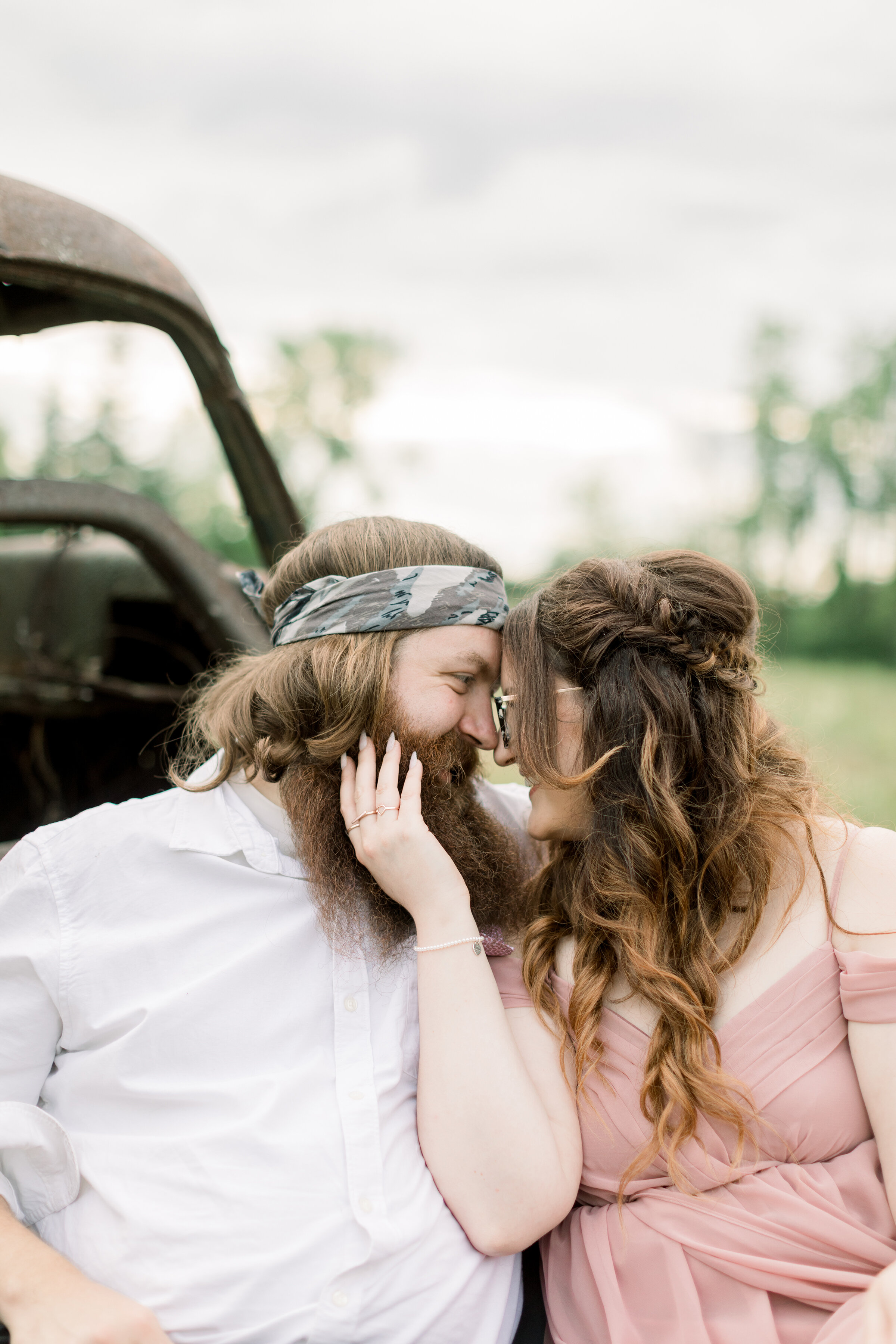  The happy couple shares a kiss during their engagement session in Ontario, Canada while sitting on a blue velvet tufted couch in a grassy field. Rusty truck vintage engagement session inspiration engagements outfit inspo camo bandana bearded groom h