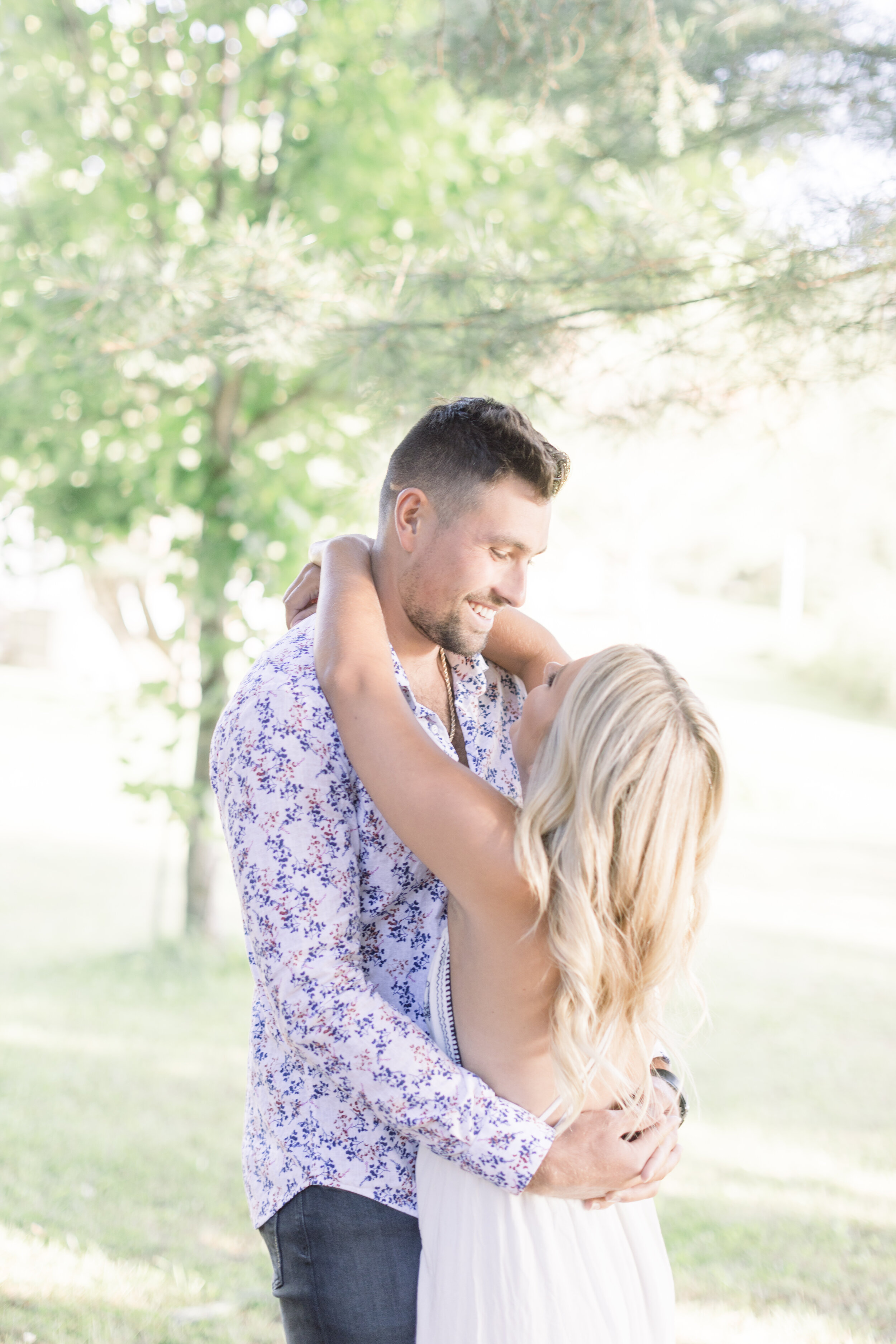  Handsome groom-to-be looks lovingly into his fiancés eyes as he wraps his arms around her waist in a loving embrace as they take their summer engagement photos in a grassy field. Summer engagement photos summer engagement photo outfit inspo sunset e
