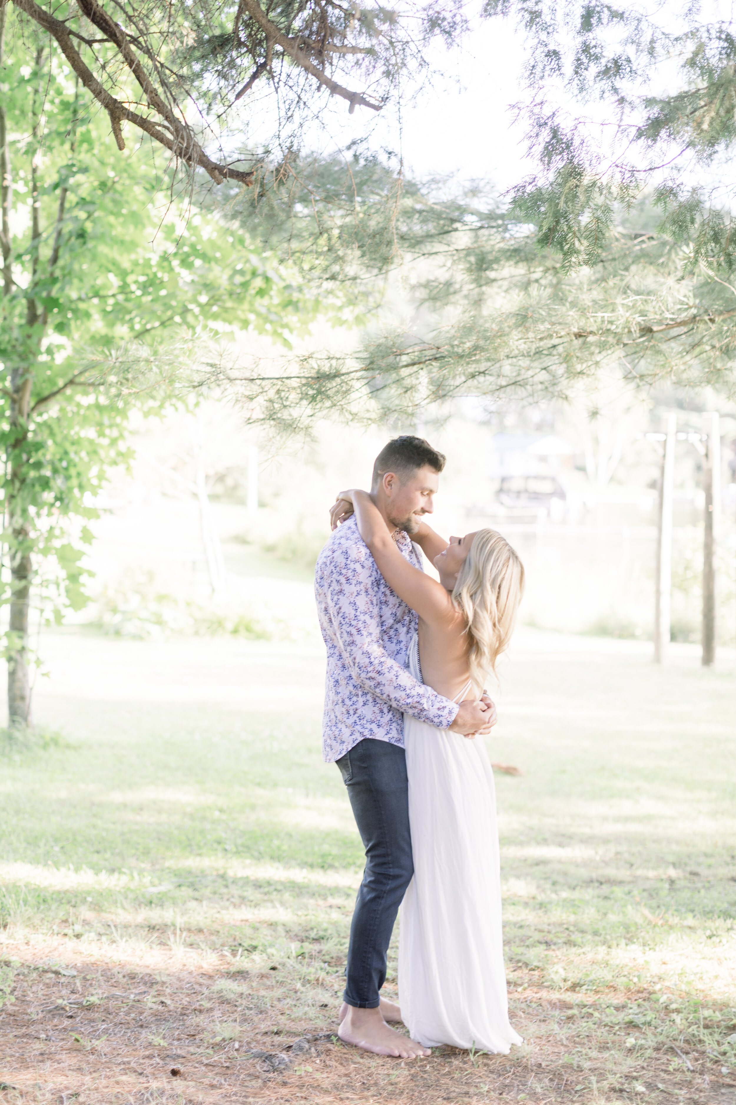  Handsome barefoot man with his arms wrapped lovingly around his fiancé in a white boho summer dress for a quebec summer engagement photography session. Professional photographer in quebec boho dress for engagement session summer sunset engagement se