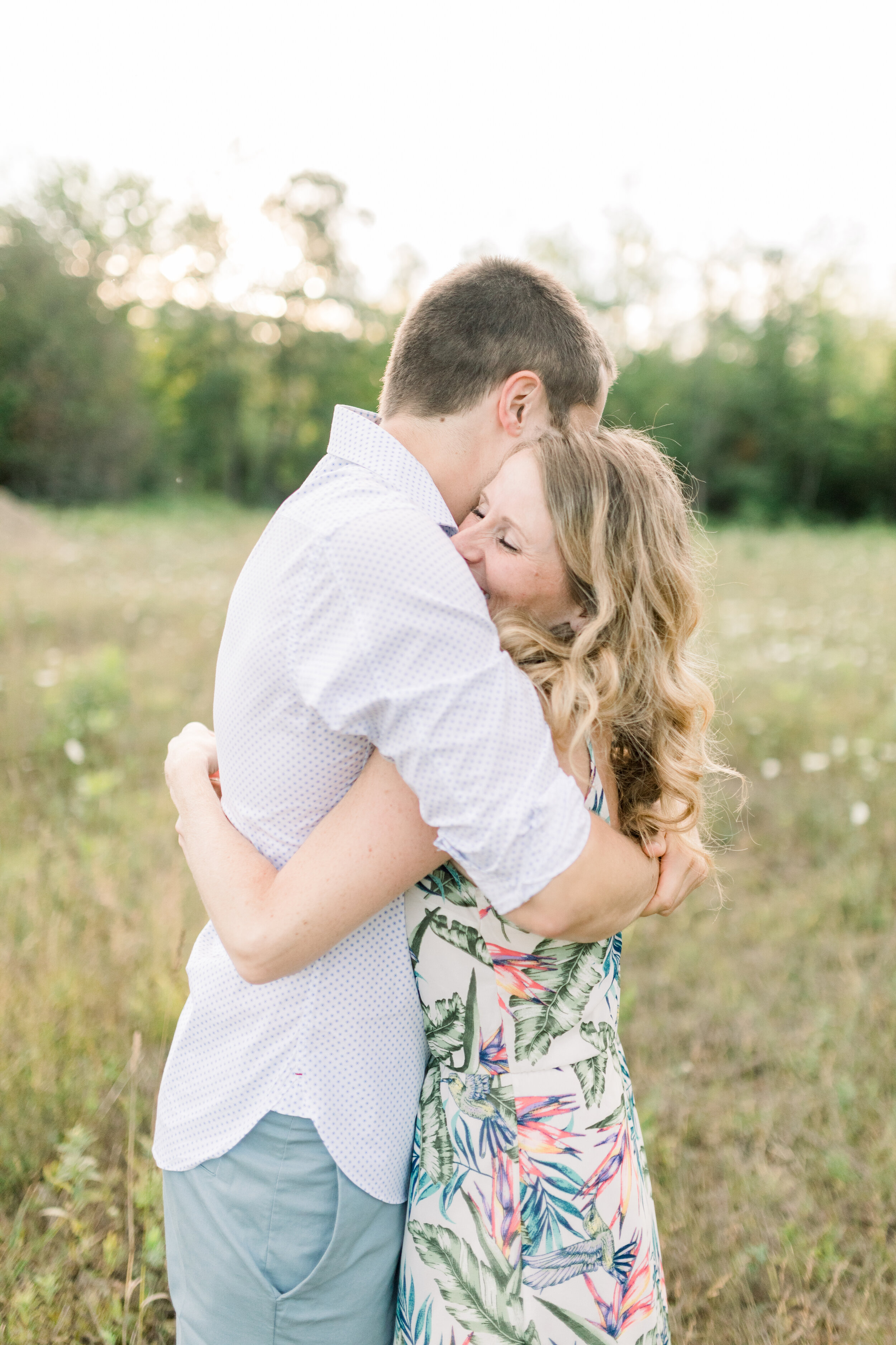  At this wildflower engagement session in Ottawa, Canada, Chelsea Mason Photography captures a candid romantic moment between this engaged couple. engaged couple hugging, womens long dirty blonde curled hairstyle, mens rolled long sleeve button up sh