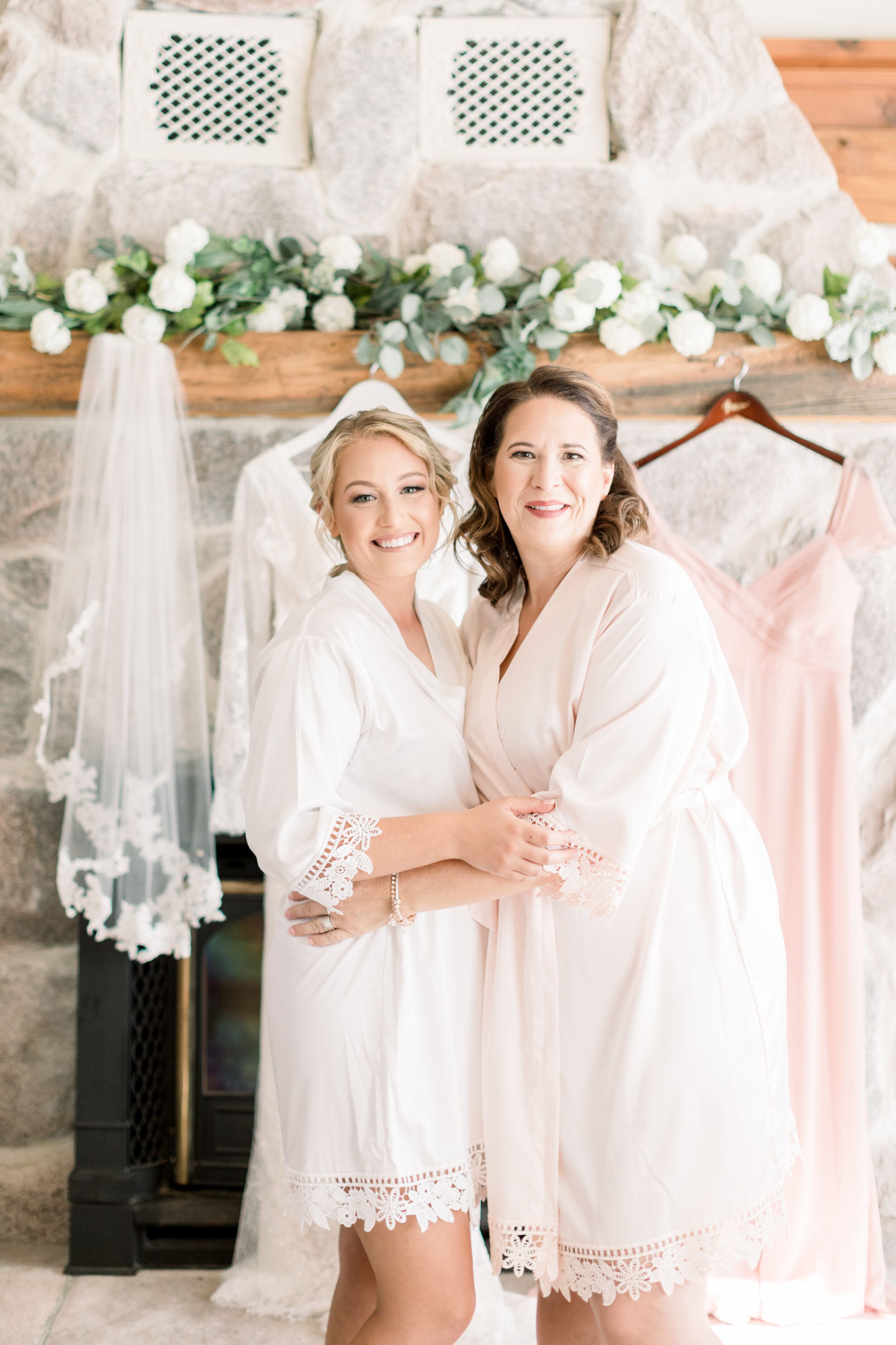  Ottowa bride with her mom standing in cute bridal robes with a lace fringe in front of a romantic fireplace lined with a strand of white and green florals. Wedding veil inspo bridal robes ottowa bride and her mom getting ready wedding photography st