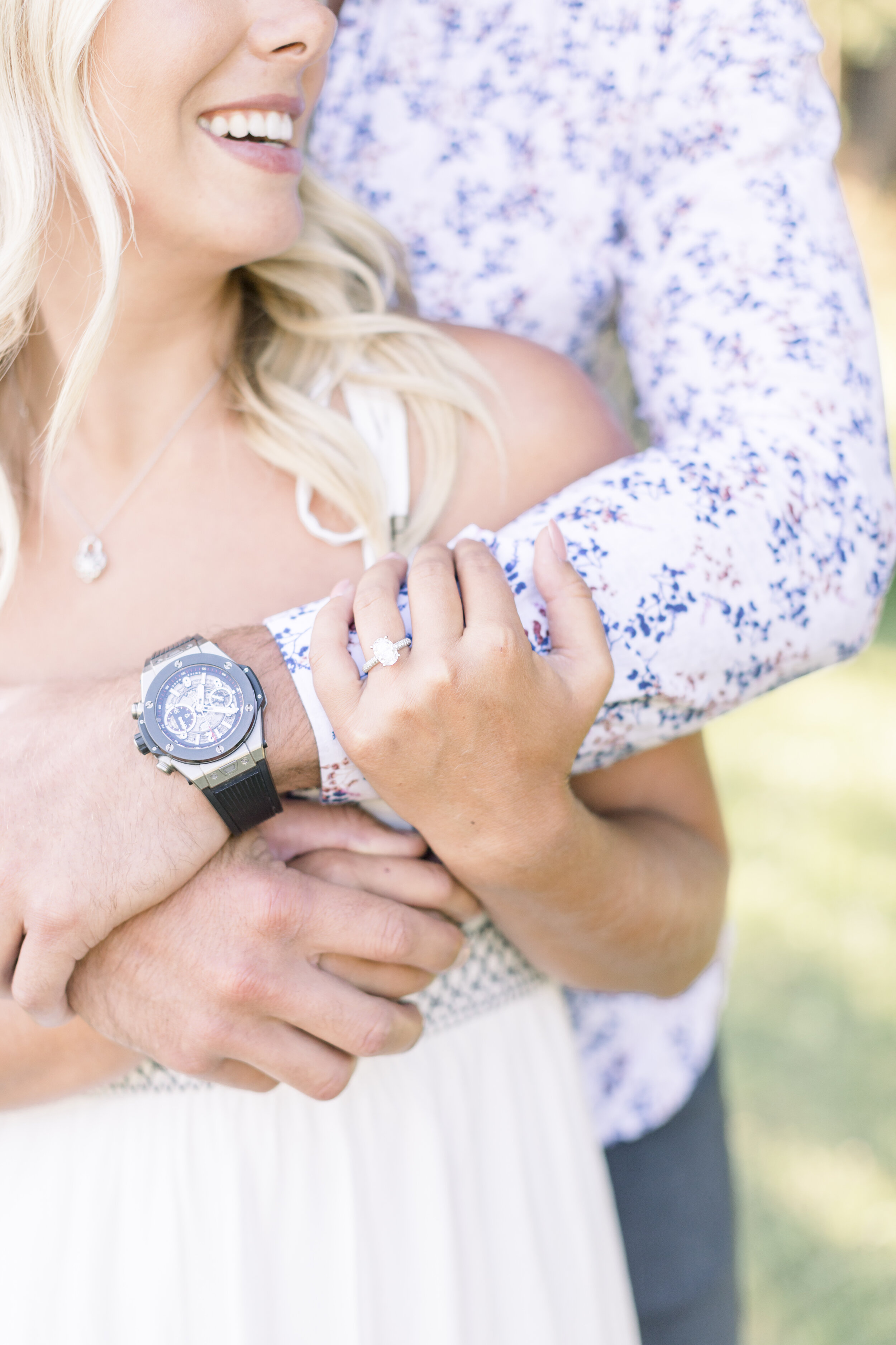  Close up detail shot of a stunning oval shaped engagement ring on a beautiful fiancé with her lovers watch-clad arms wrap around her for a summer cottage engagement session at sunset. Engagement photography session at sunset in quebec photographer f