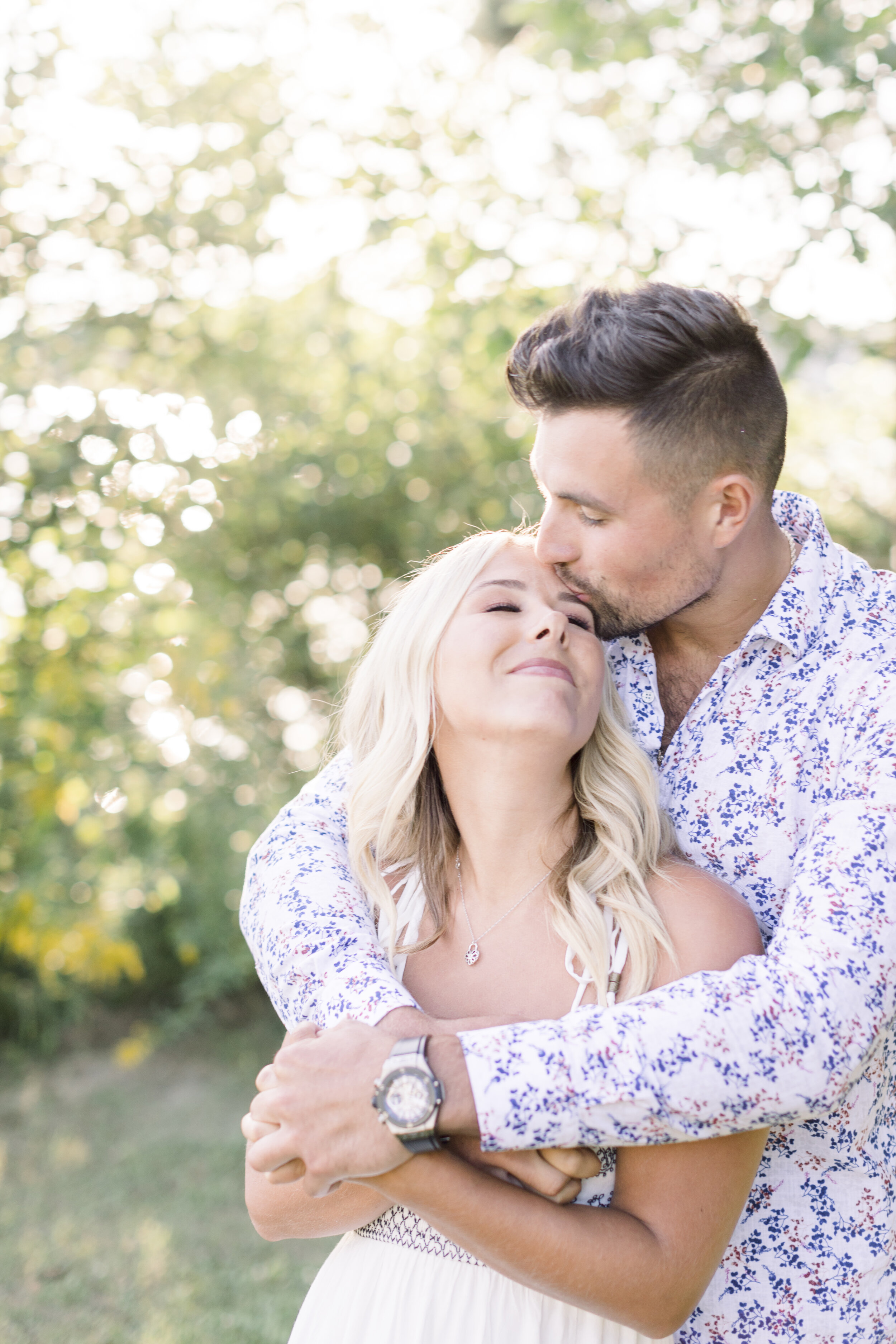  Handsome man wraps his arms around his beautiful fiancés dressed in a summer boho outfit as he kisses her forehead in the meadow for their summer sunset engagement session in quebec. Forehead kisses during engagement sunset photography session white