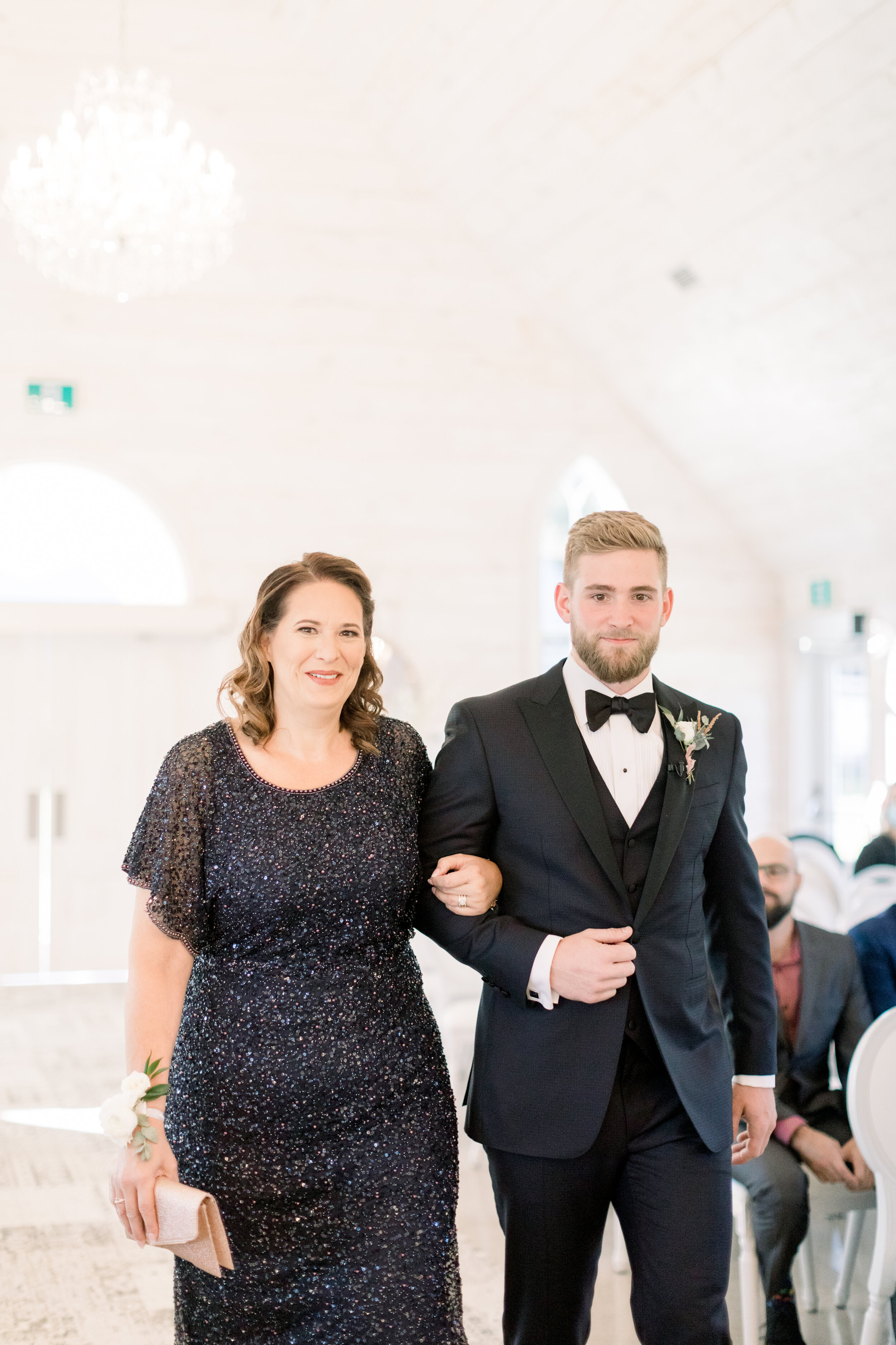  Handsome groom in a boutineer and black bowtie escourting his mom dressed in a shiny black dress down the wedding ceremony aisle at stonefield venue in ottowa. Groom in a bowtie groom escourting his mom wedding ceremony processional ottowa wedding p