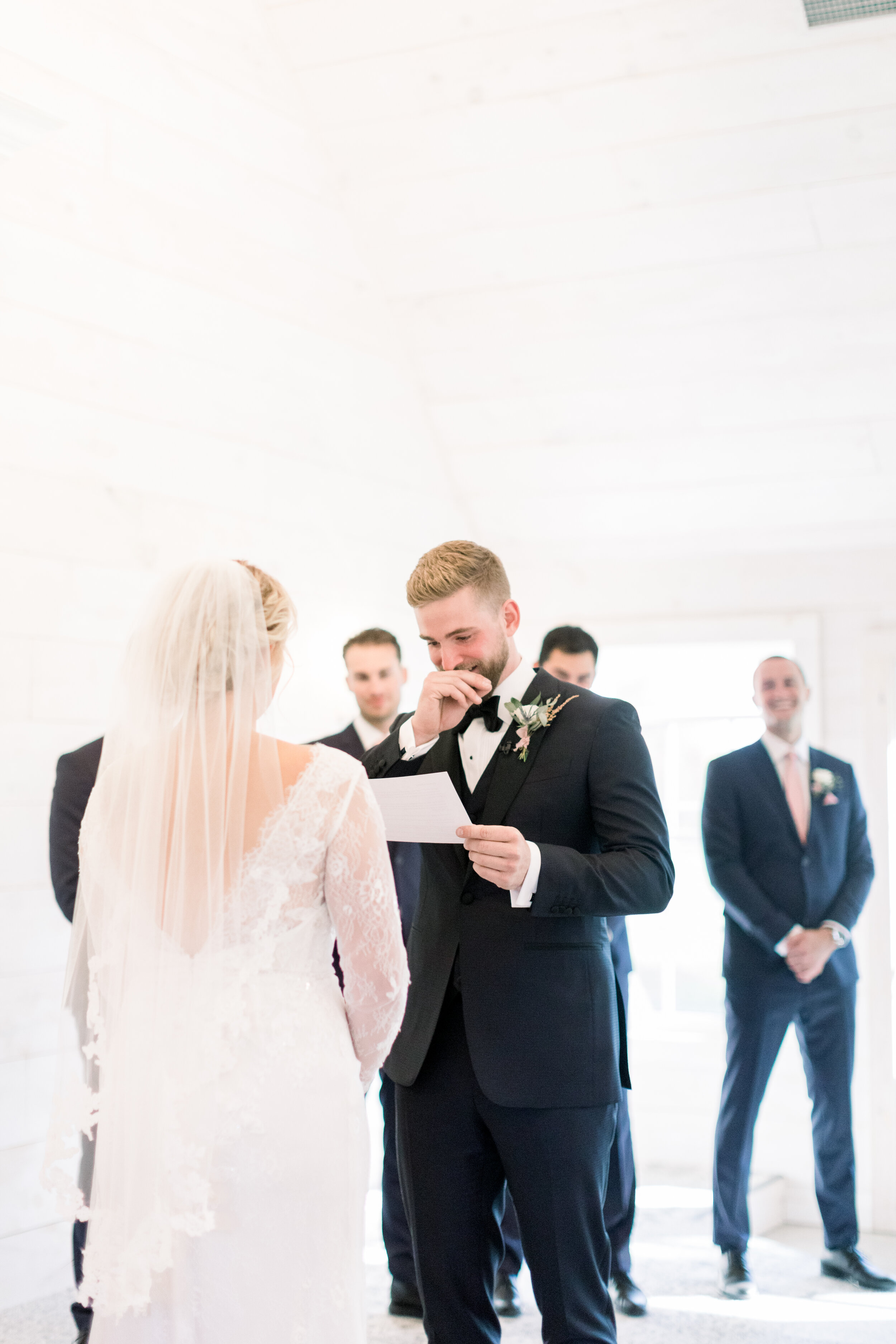  Handsome groom in a black bowtie getting emotional as he reads his wedding vows to his blushing bride in a stunning white wedding dress and a midlength lace trimmed veil at stonefield wedding venue. Wedding vows emotional groom reading vows intimate