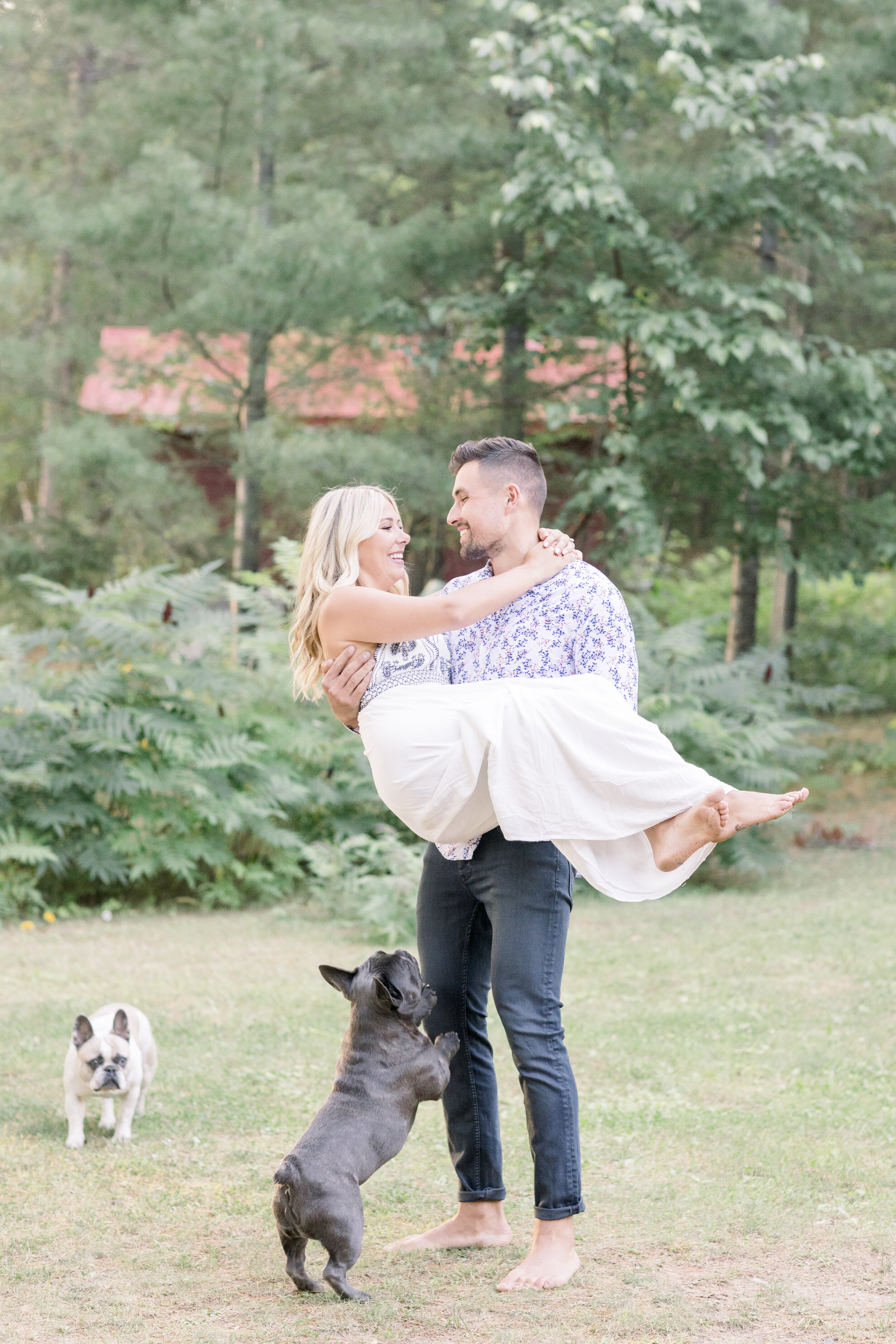  handsome man whisks his fiancé off her feet as they stand barefoot in a field outside a summer cottage in quebec as their frenchie pups run around them during their sunset engagement photography session. Sunset engagement session engagement session 