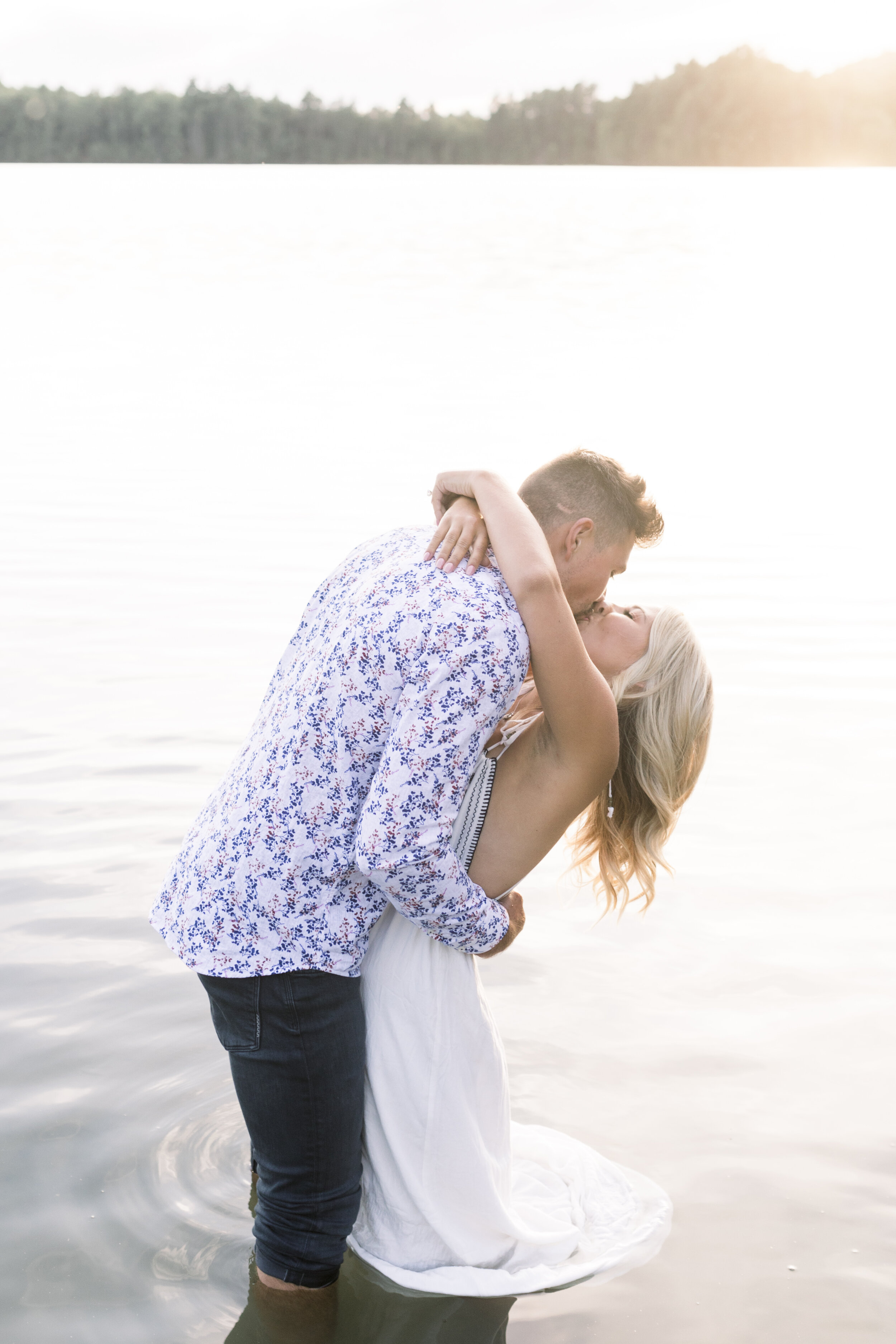  Handsome man dips his fiancé in a kiss as they stand in the lake for their sunset engagement session in Lac Mallonne, quebec. Quebec engagement session lakeside engagement photography engagement sessions in the water engagement sessions in a lake qu