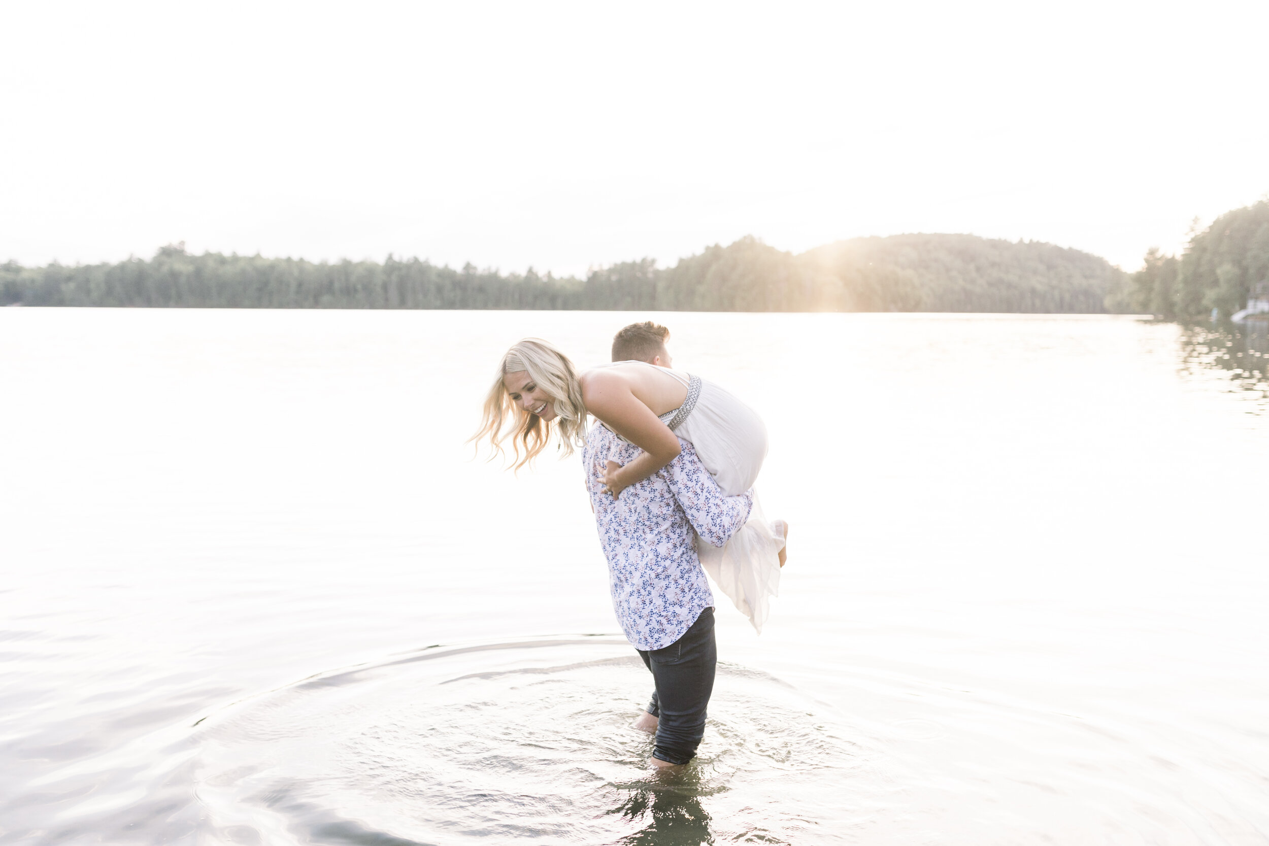  Handsome man playfully picks up his fiancé and throws her over his should as they take engagement photos in the lake at a quebec cottage at sunset. Playful engagement photography poses photography session at the lake engagement photography in quebec