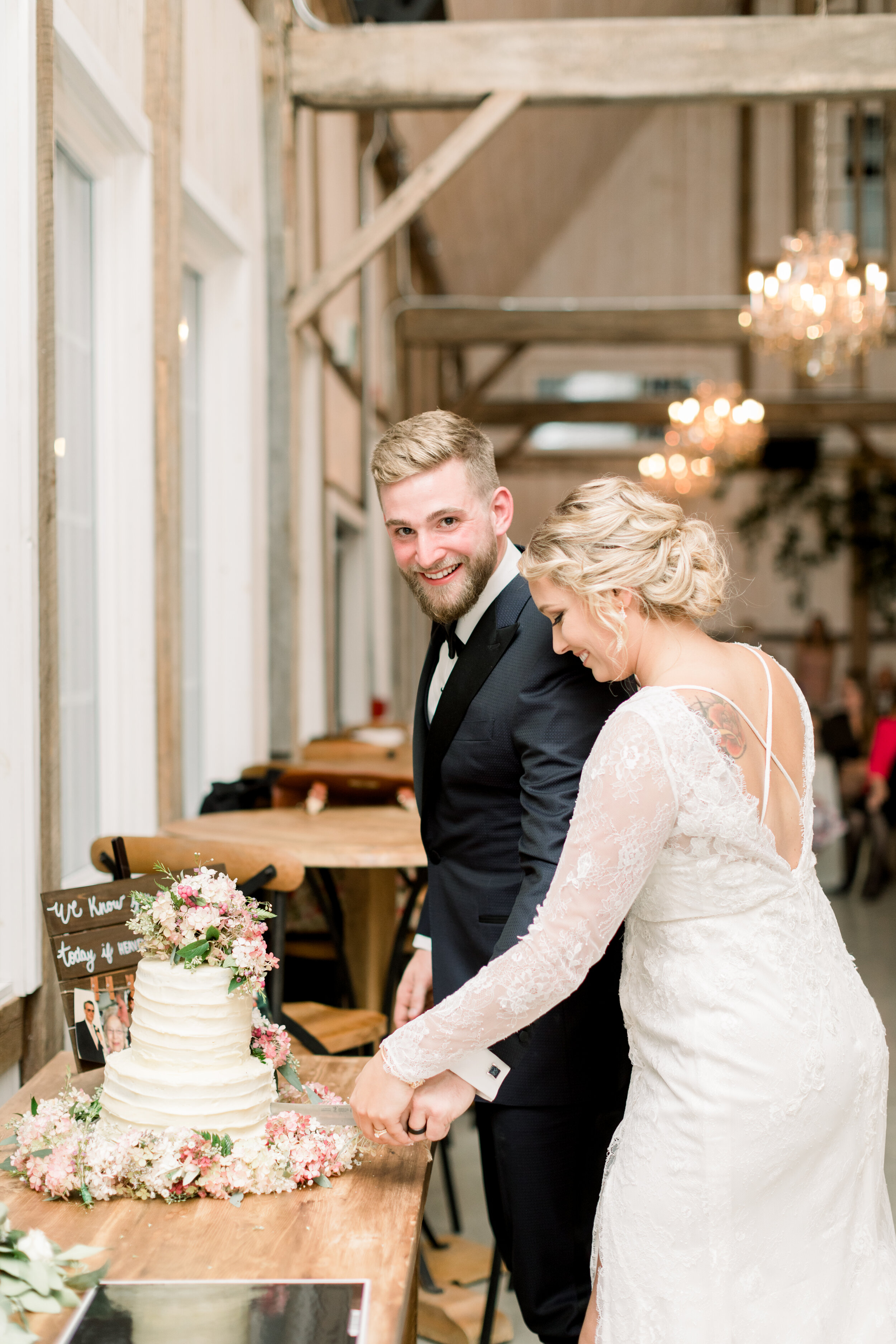  Tattoed Ottowa bride with a crisscross back lace wedding dress and a beautiful updo standing with her groom cutting a rustic cake with cascading pink florals. Rustic wedding cake wedding cake with florals tiered wedding cake inspo ottowa bride and g