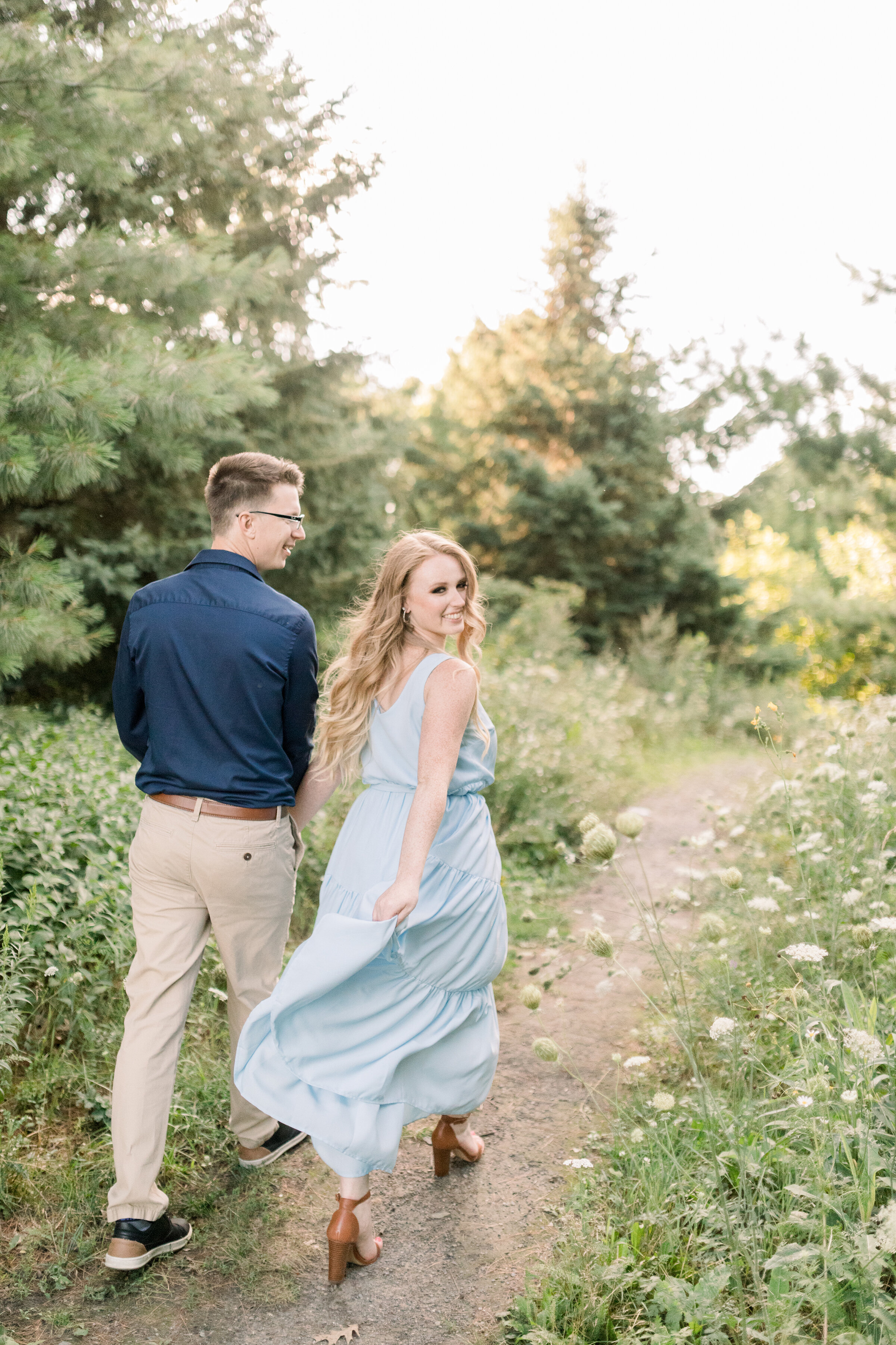  A glowing soon to be bride looks back at the camera as she’s lead by her fiancé in a beautiful Dominion Arboretum engagement photo shoot in Ottawa by Chelsea Mason Photography. Couple walking pose inspiration couple goals engagement photo shoot insp