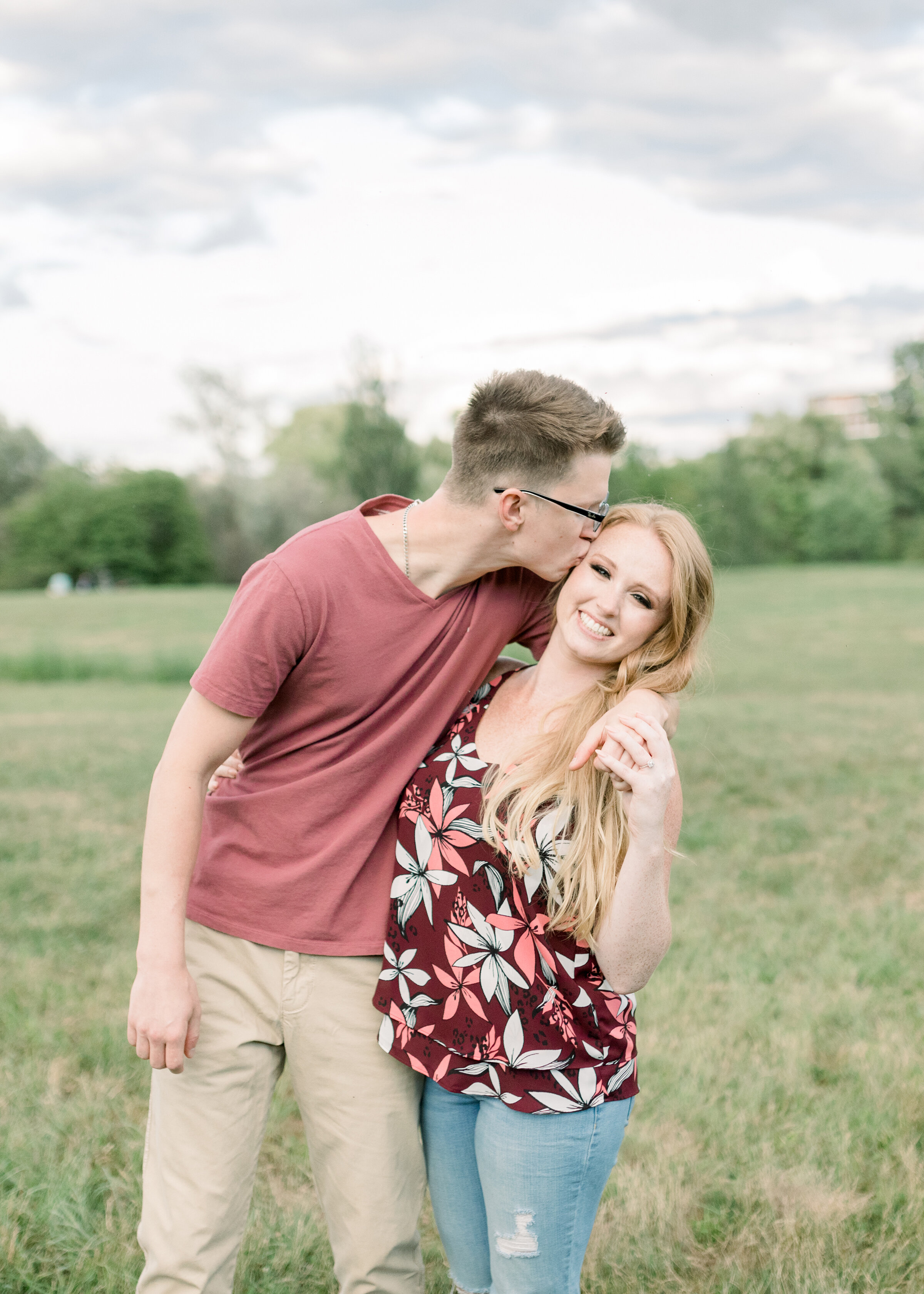  A soon to be groom kisses his fiancé on the cheek as she smiles at the camera in an engagement styled photo shoot in Dominion Arboretum, Ottawa. Casual engagement session attire for women casual attire for men  client attire inspiration engagement s