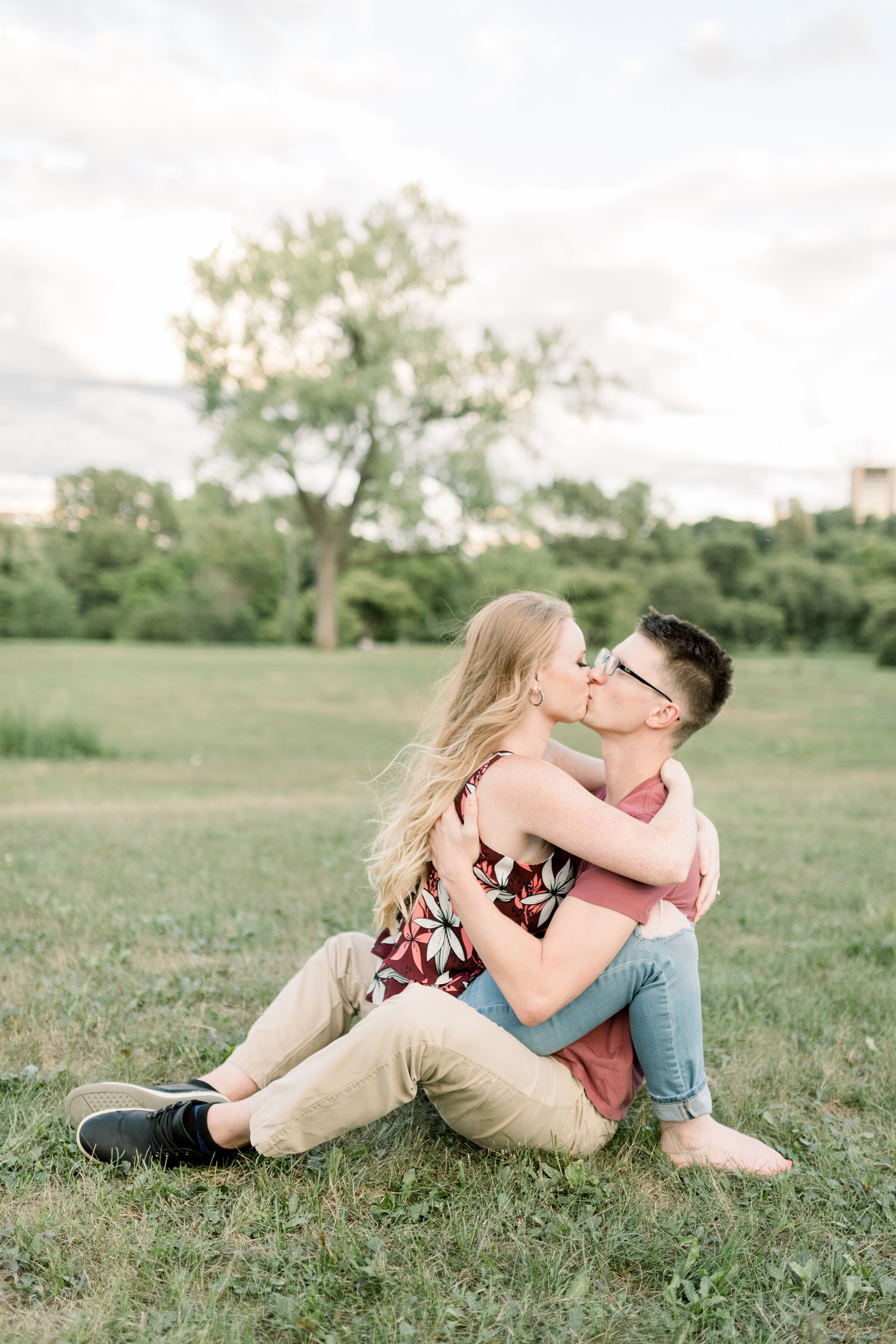  A glowing soon to be bride sits on her fiancés lap as they kiss in a romantic Dominion Arboretum styled engagement session in Ottawa. Professional engagement photographer Chelsea Mason Photography couple sitting pose inspiration ideas and goals kiss