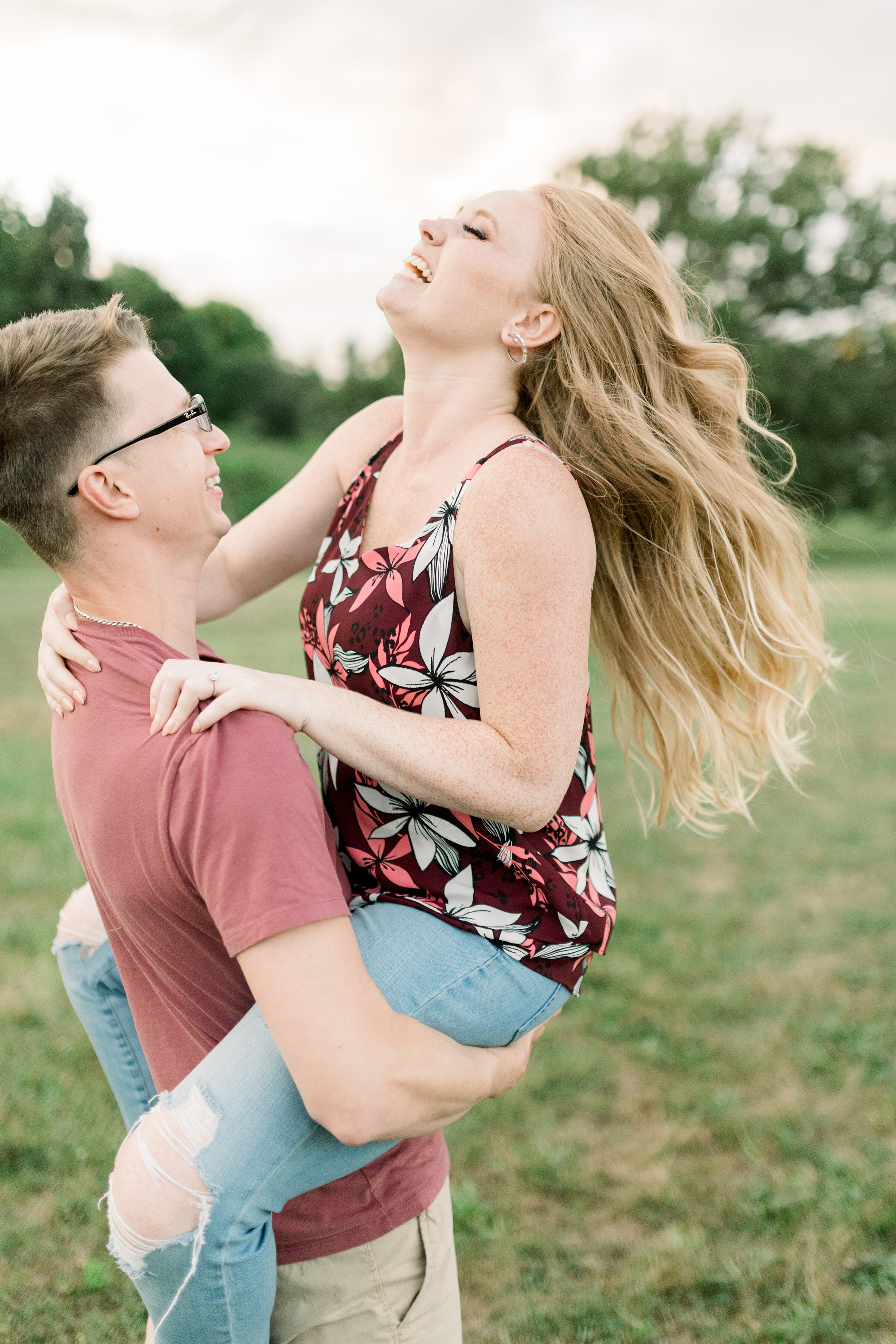  A glowing woman laughs as her fiancé lifts her off the ground in a playful engagement session at Dominion Arboretum in Ottawa by professional engagement photographer Chelsea Mason Photography. Engagement session couple pose inspiration ideas and goa