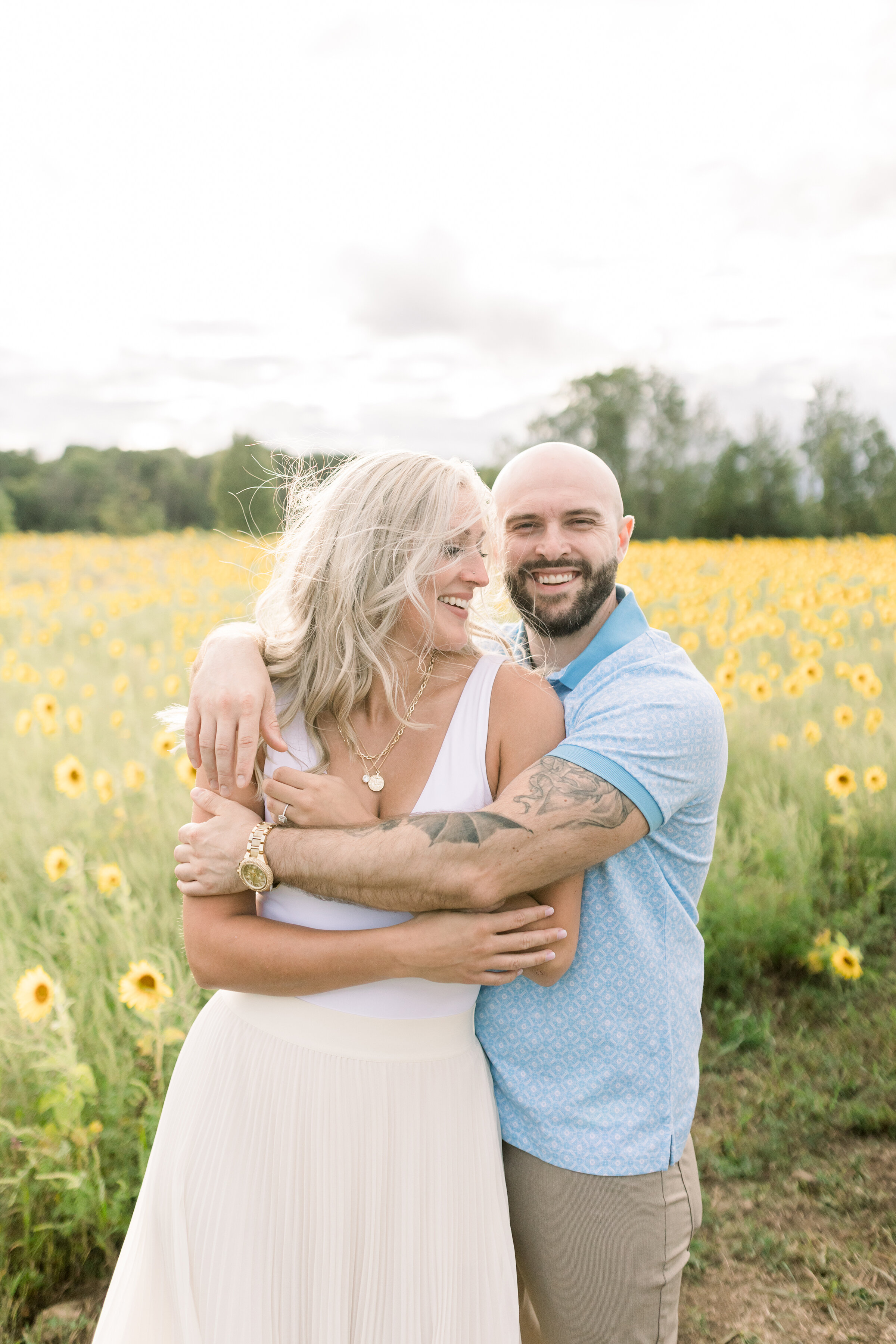  A glowing couple laugh and hold each other as they stand in an Ottawa, Ontario sunflower field in a unique engagement session by professional engagement photographer Chelsea Mason Photography. Couple pose inspiration ideas and goals summer client at