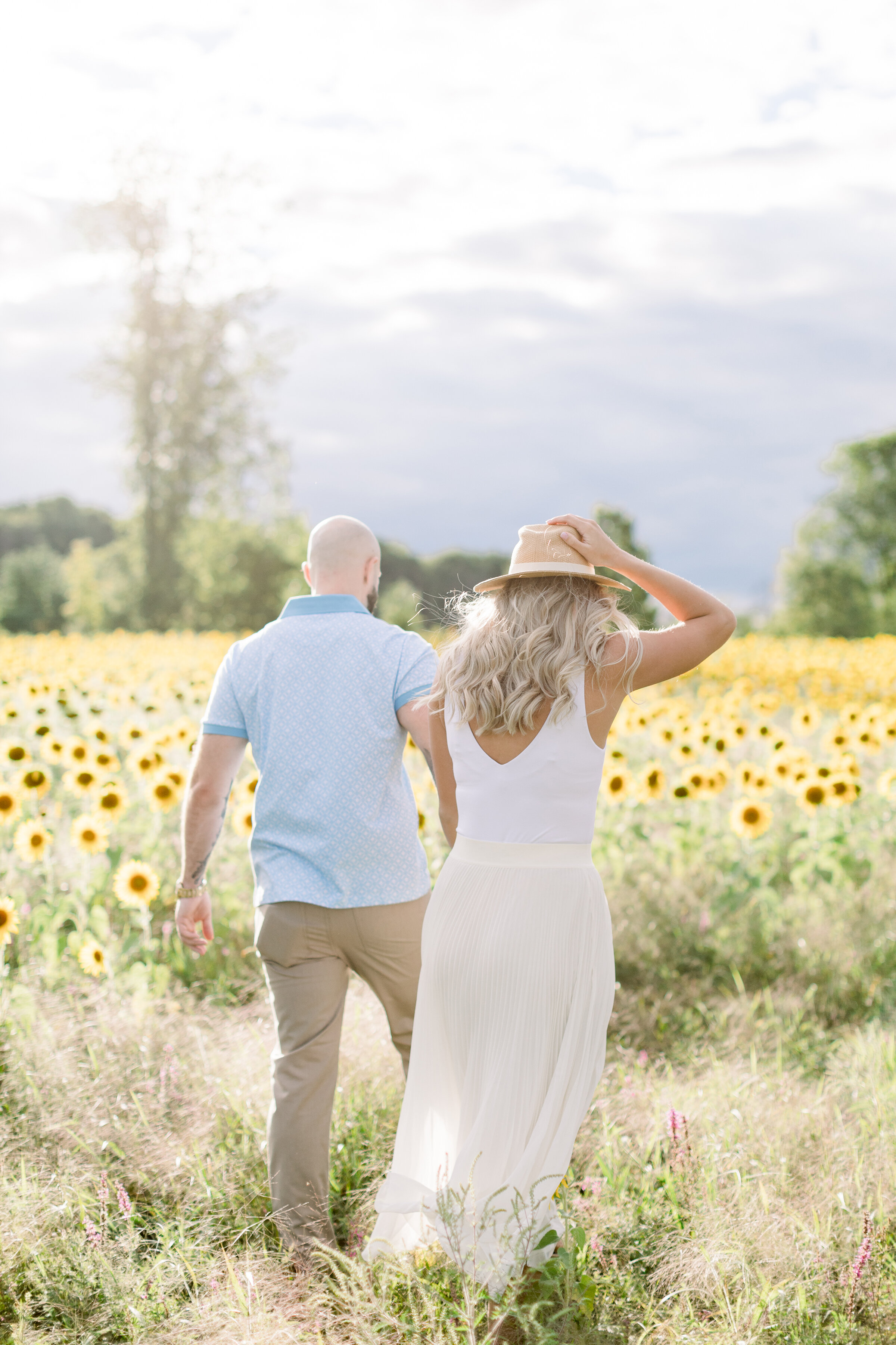  A beautiful couple walk through a field of sunflowers as she holds her hat in a sunflower styled engagement session in Ottawa, Ontario. Professional engagement photographer Chelsea Mason Photography walking couple pose inspiration ideas and goals su