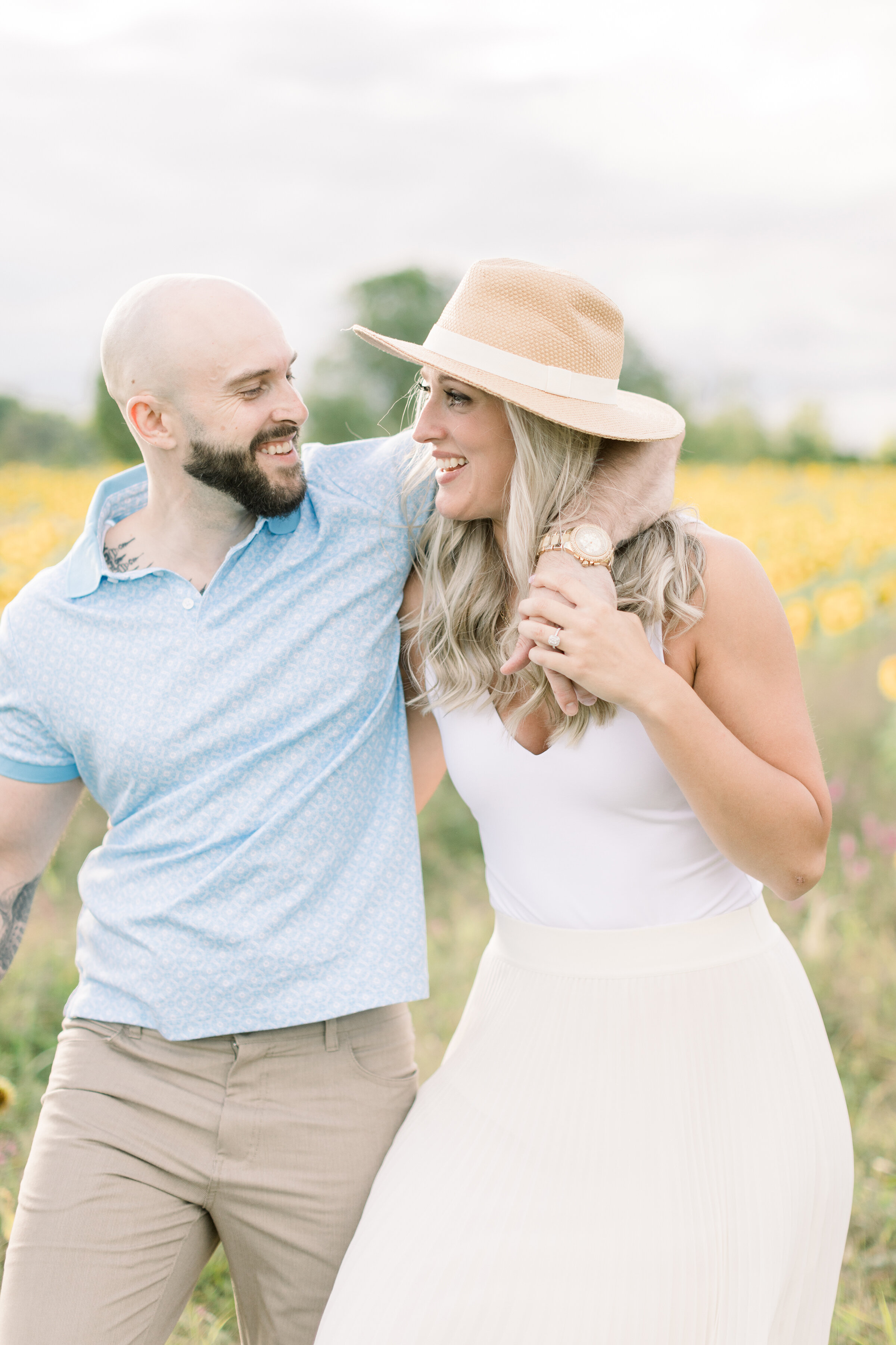  Soon to be bride and groom smile at each other as they walk in a beautiful warm sunflower engagement session in Ashton, Ontario by professional photographer Chelsea Mason Photography. Summer goals summer engagement outfit inspiration ideas and goals