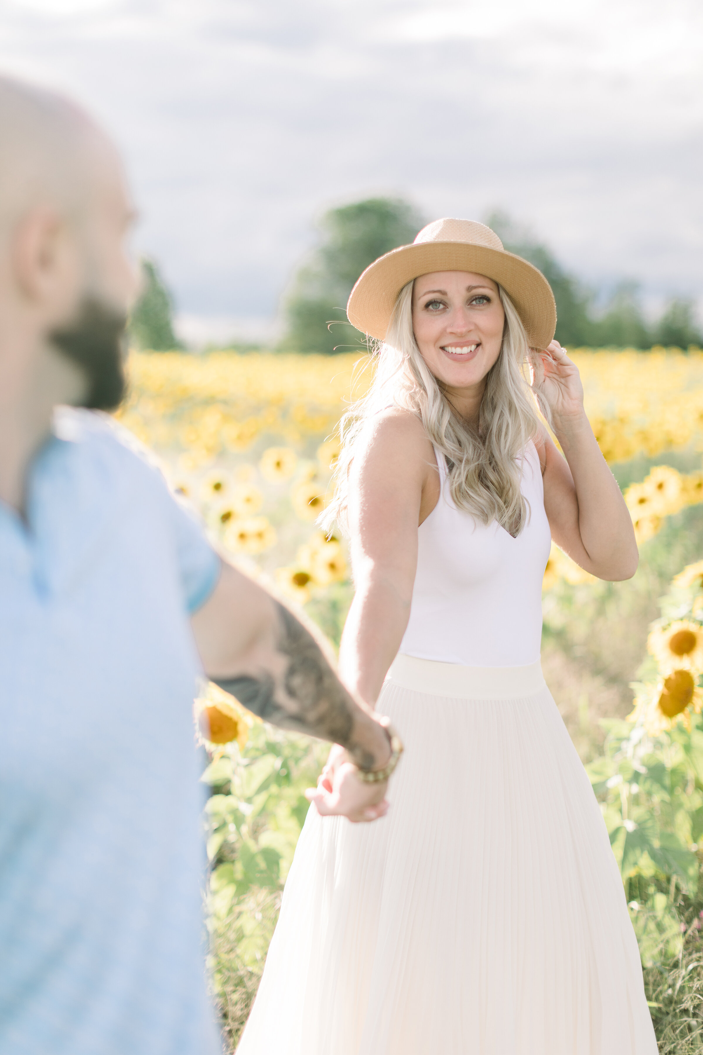  The beautiful soon to be bride holds her fiancés hand as she smiles at the camera in a bright and airy summer engagement session in Ashton, Ontario’s sunflower field. Couple goals professional engagement photographer Chelsea Mason Photography outdoo