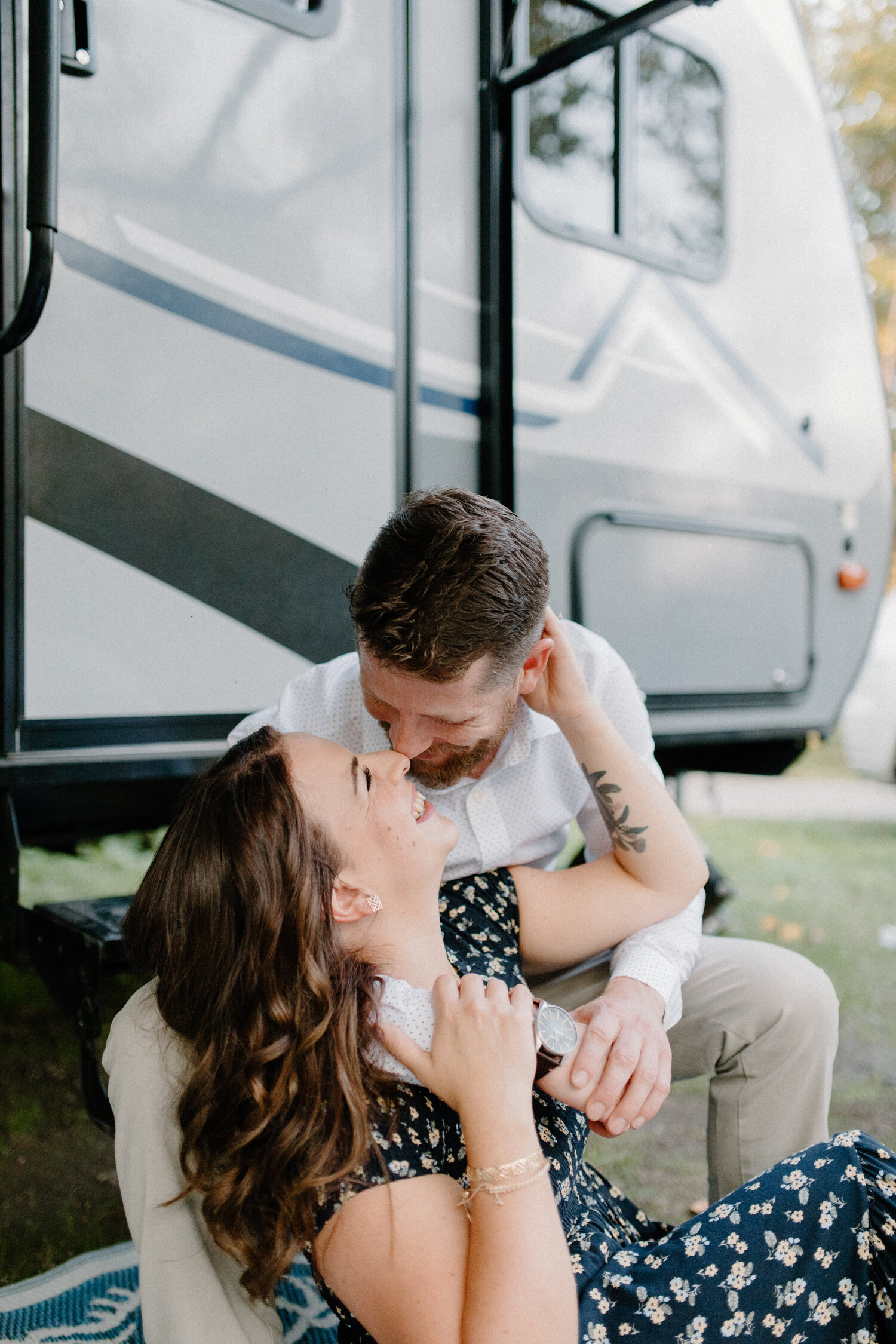  While sitting on a picnic blanket positioned outside their camper rv, Chelsea Mason Photography captures this groom kissing his bride and smiling during this engagement session in the woods near Ottawa, Canada. Wooded outdoorsy camper engagement ses