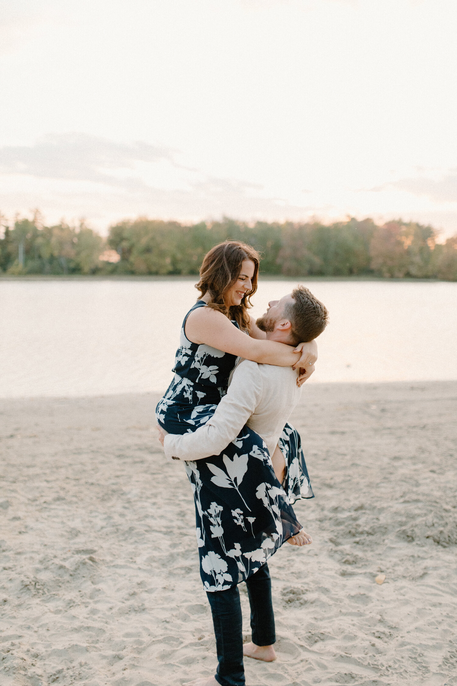  This playful soon-to-be groom picks up his finance and hold her in his arms during this beach engagement session in Ottawa, Canada by Chelsea Mason Photography. Formal beach engagement session outfit, man holding woman in his arms and laughing, play