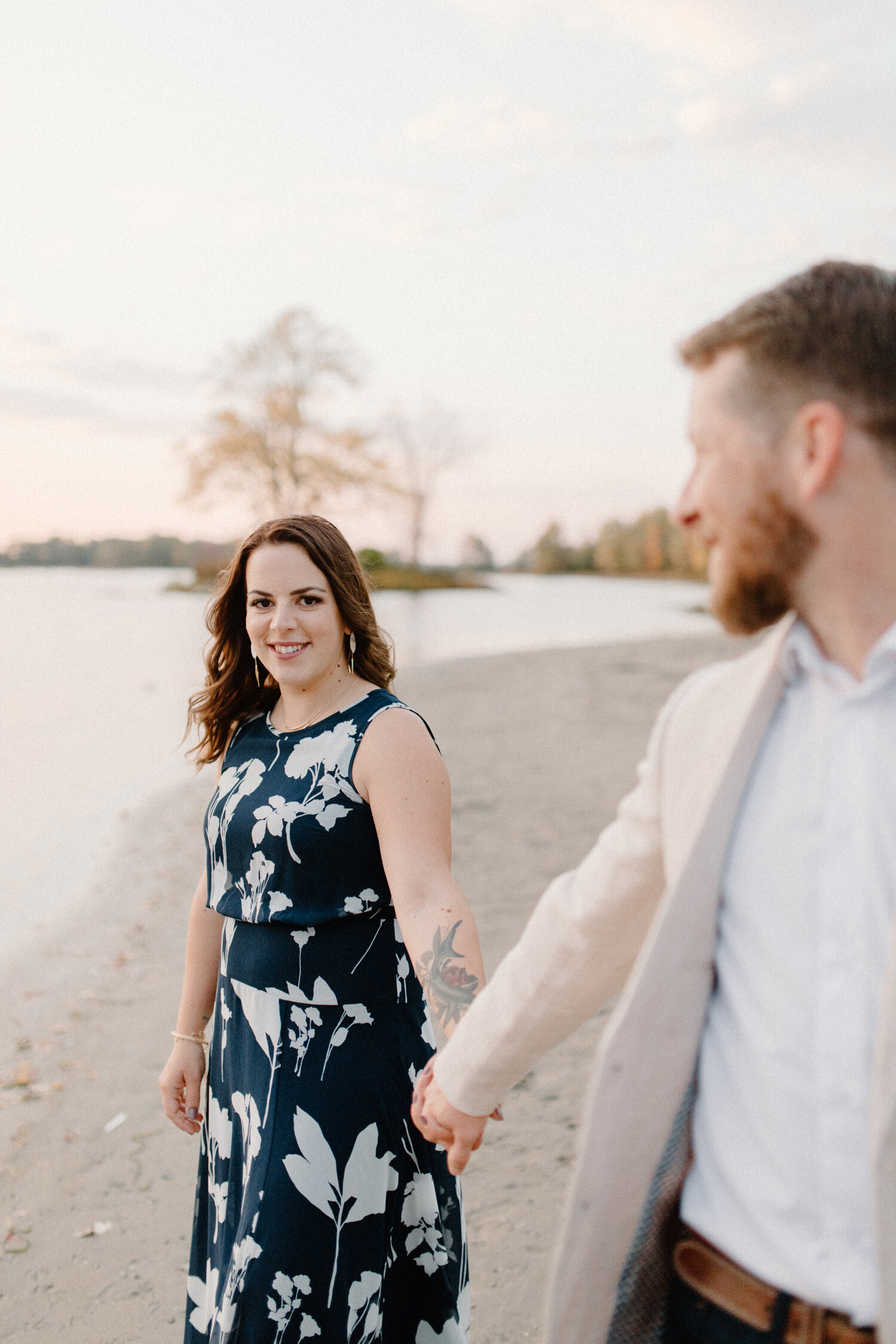  Ontario, Canada engagement photographer, Chelsea Mason Photography captures this engaged couple holding hands while walking along the beach shore. Womens floral forearm colored tattoo, womens high low black and white large print maxi dress, formal b