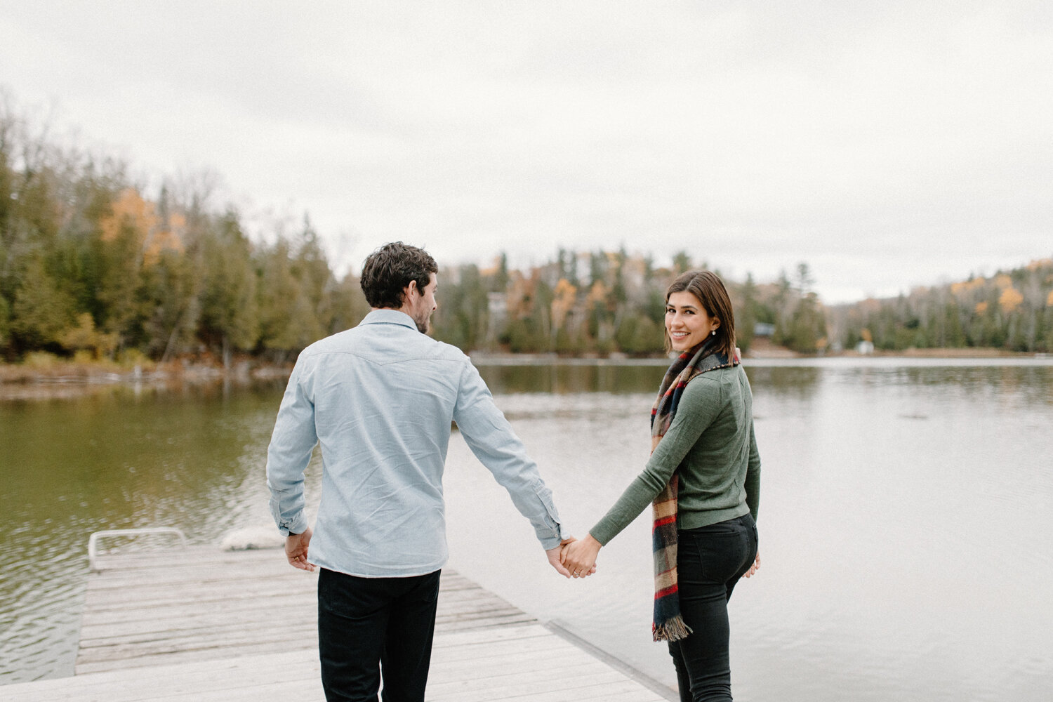  During this autumn engagement session in Quebec, Canada, Chelsea Mason Photography captures this engaged couple holding hands while smiling and walking on a pier on a moody overcast day. Moody overcast engagement session, couple walking hand-in-hand