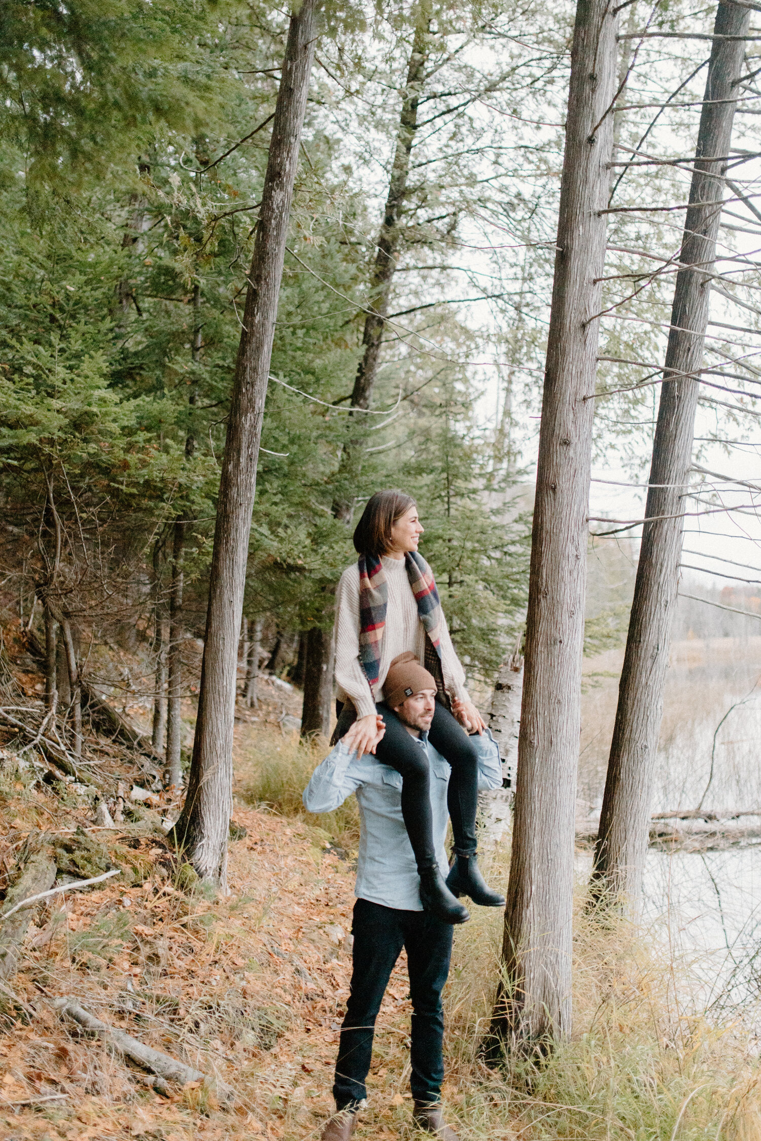  On the wooded shoreline of this Quebec, Canada lake, Chelsea Mason Photography captures a playful engagement session where this woman sits on her fiance’s shoulders. Woman sitting on fiance’s shoulders, playful piggy back ride, overcast autumn engag