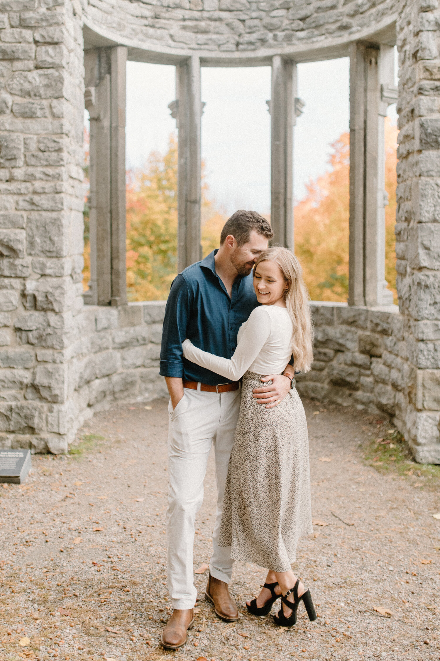  Chelsea Mason Photography captures this engaged couple hugging and smiling romantically at one another during their engagement session in a aged brick gazebo. autumn quebec canada engagement session, engaged couple tenderly hugging, womens black str
