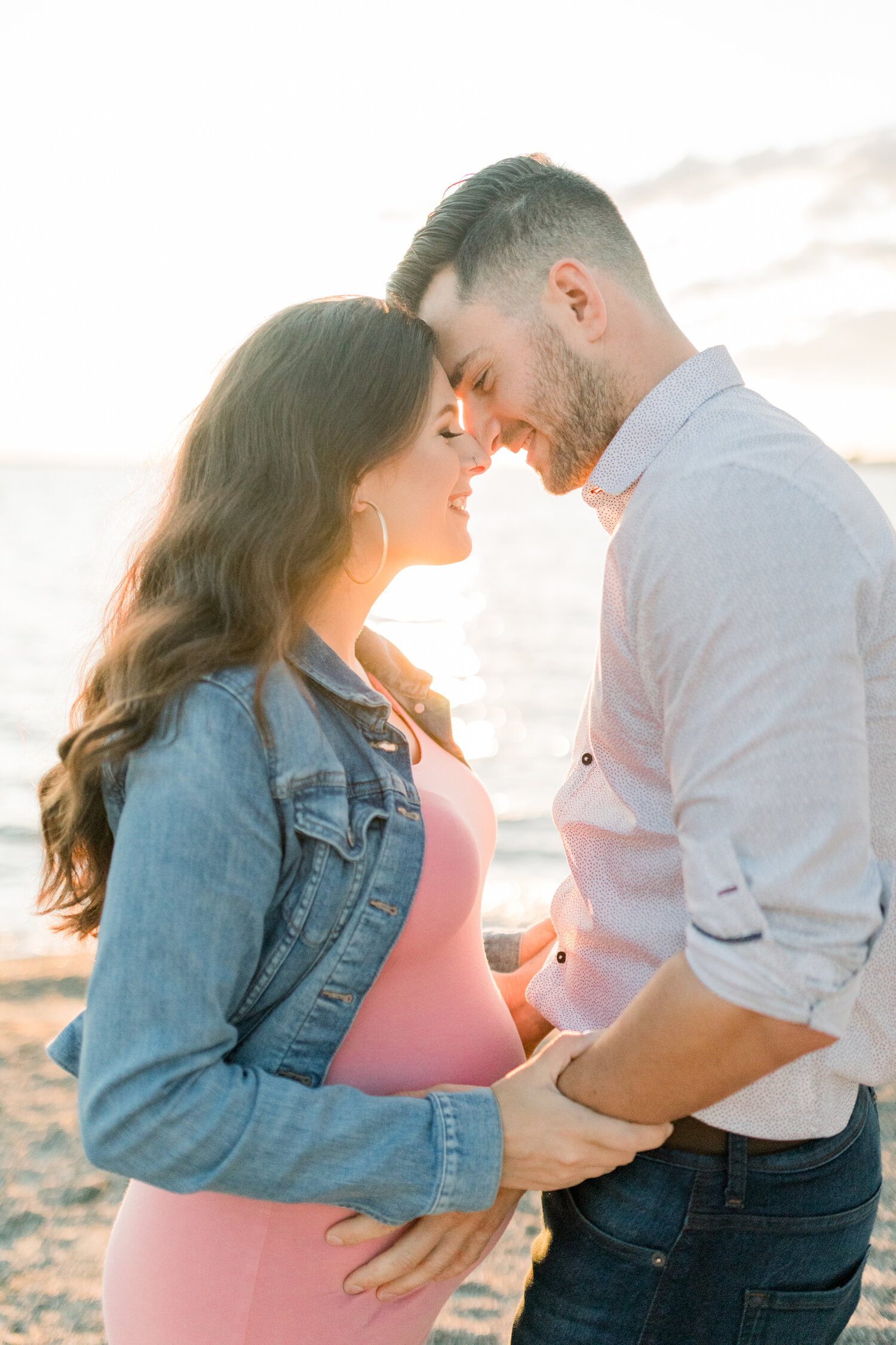  Along the shoreline of Brittania beach in Ottawa, Canada, Chelsea Mason Photography captures this expectant father holding his wife’s stomach. fitted maternity pink dress, womens denim jacket, brittana beach, beach maternity photos, ottawa canada ma