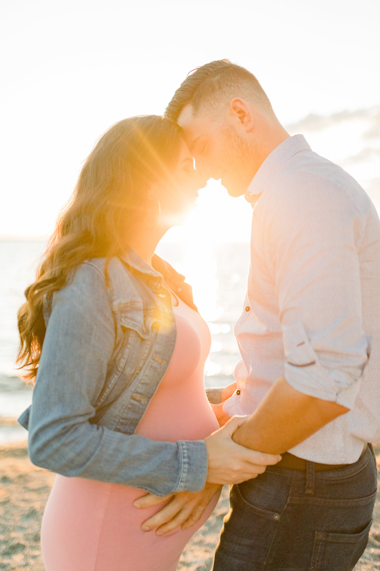  With the setting sun beaming behind them, Chelsea Mason Photography captures this expectant couple embracing along the beach in Ottawa, Canada. expectant father holding wife’s stomach, fitted maternity pink dress, brittania beach ottawa canada mater