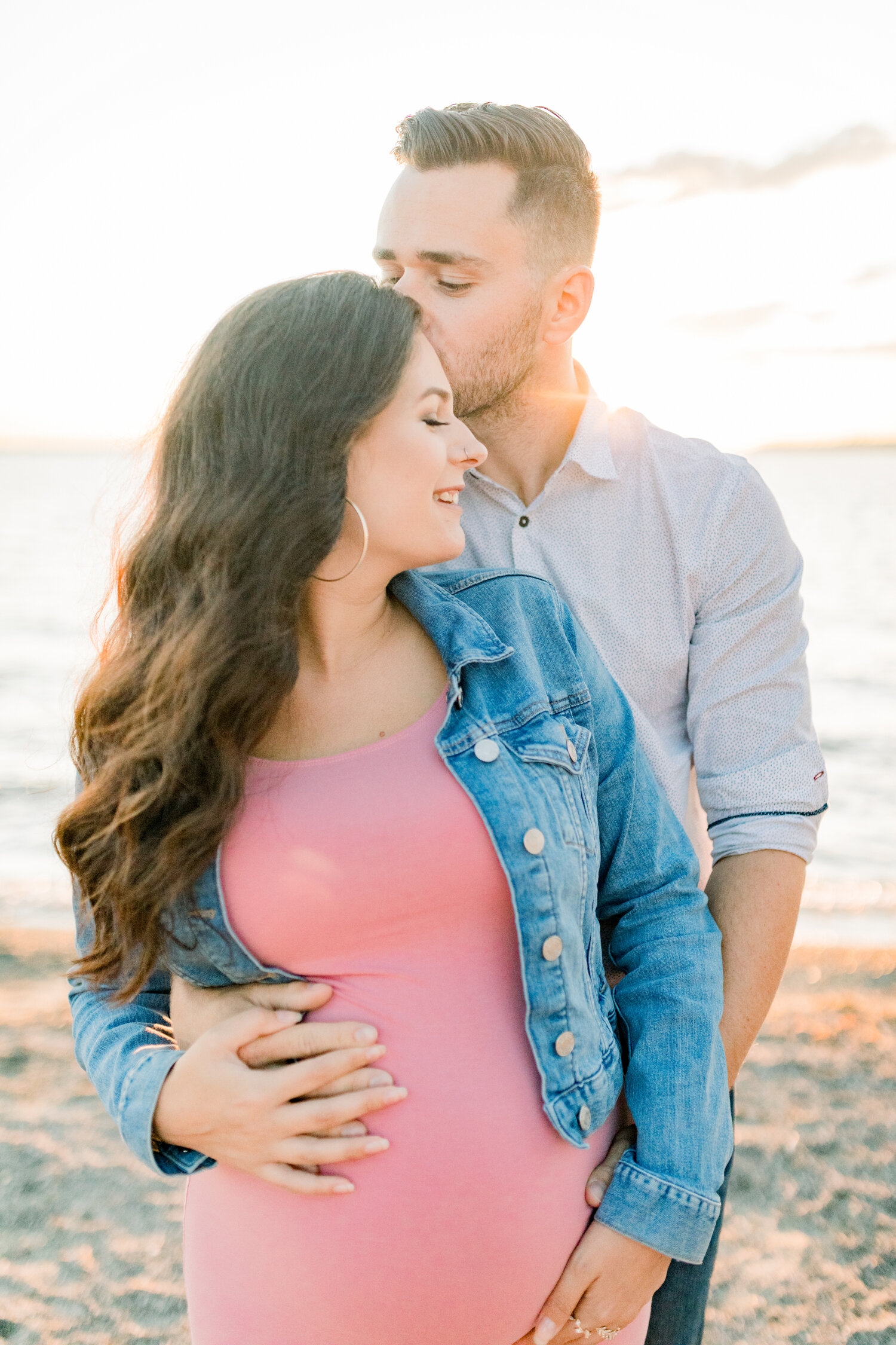  Hugging her from behind, this expectant father tenderly embraces his pregnant wife during this maternity session by Chelsea Mason Photography in Ontario, Canada. pink and denim colored family photos, long medium brown colored waved hair, womens deni
