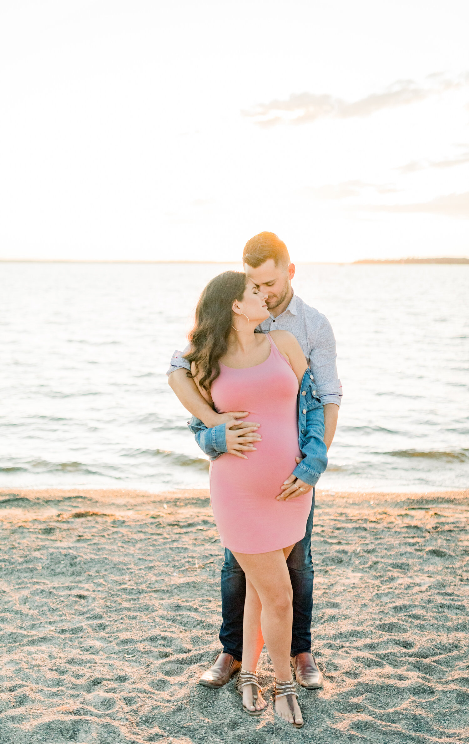  With the sun setting behind them, Ontario, Canada maternity photographer, Chelsea Mason Photography captures this expectant couple romantically embracing. womens fitted pink spaghetti strapped maternity dress, oversized denim jacket hanging off the 