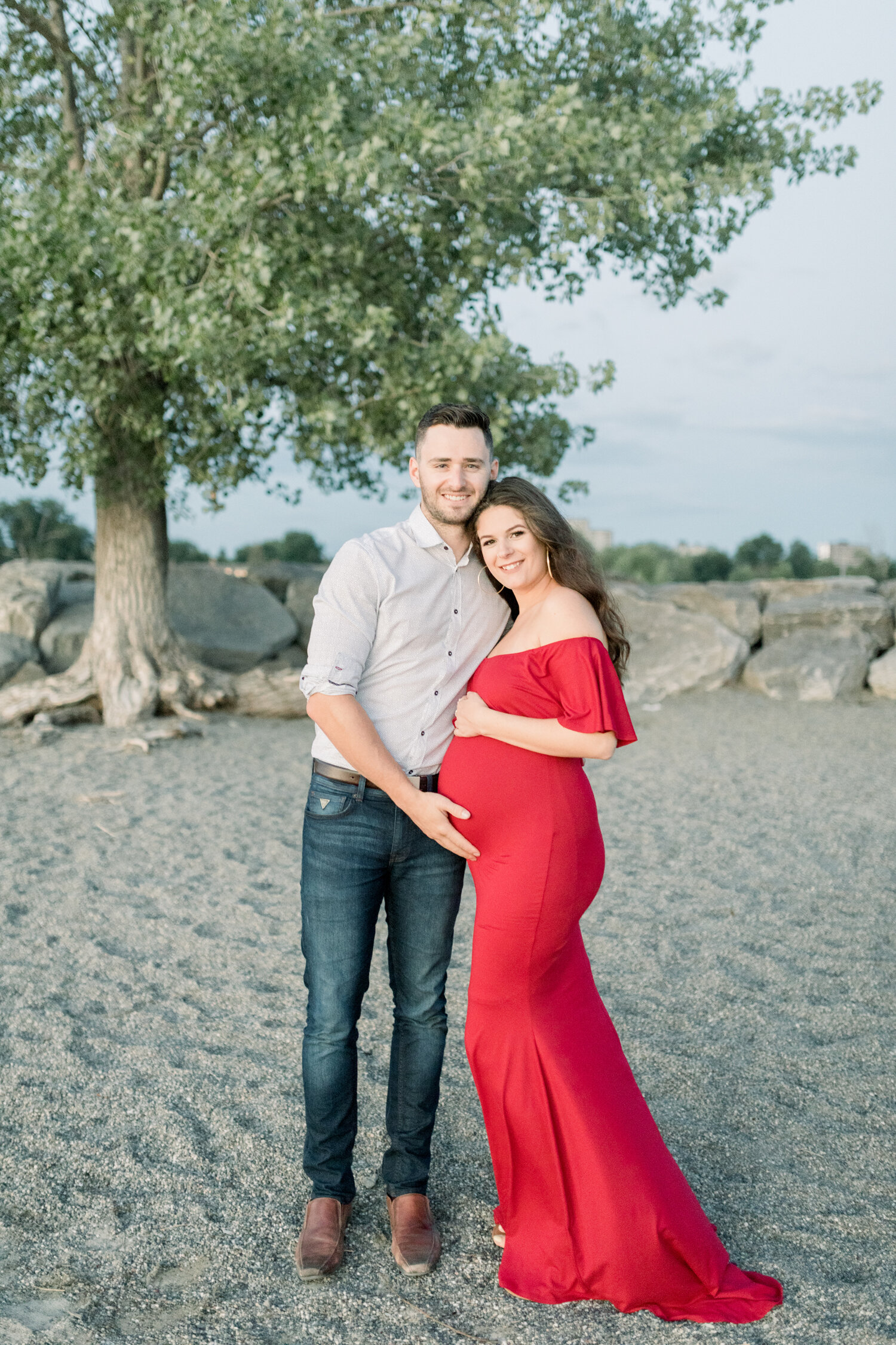  With boulders and shade trees in the backdrop, this expectant couple poses during their maternity session with Chelsea Mason Photography along the sandy Brittania Beach in Ontario, Canada. ontario canada maternity photos, womens maternity fitted off