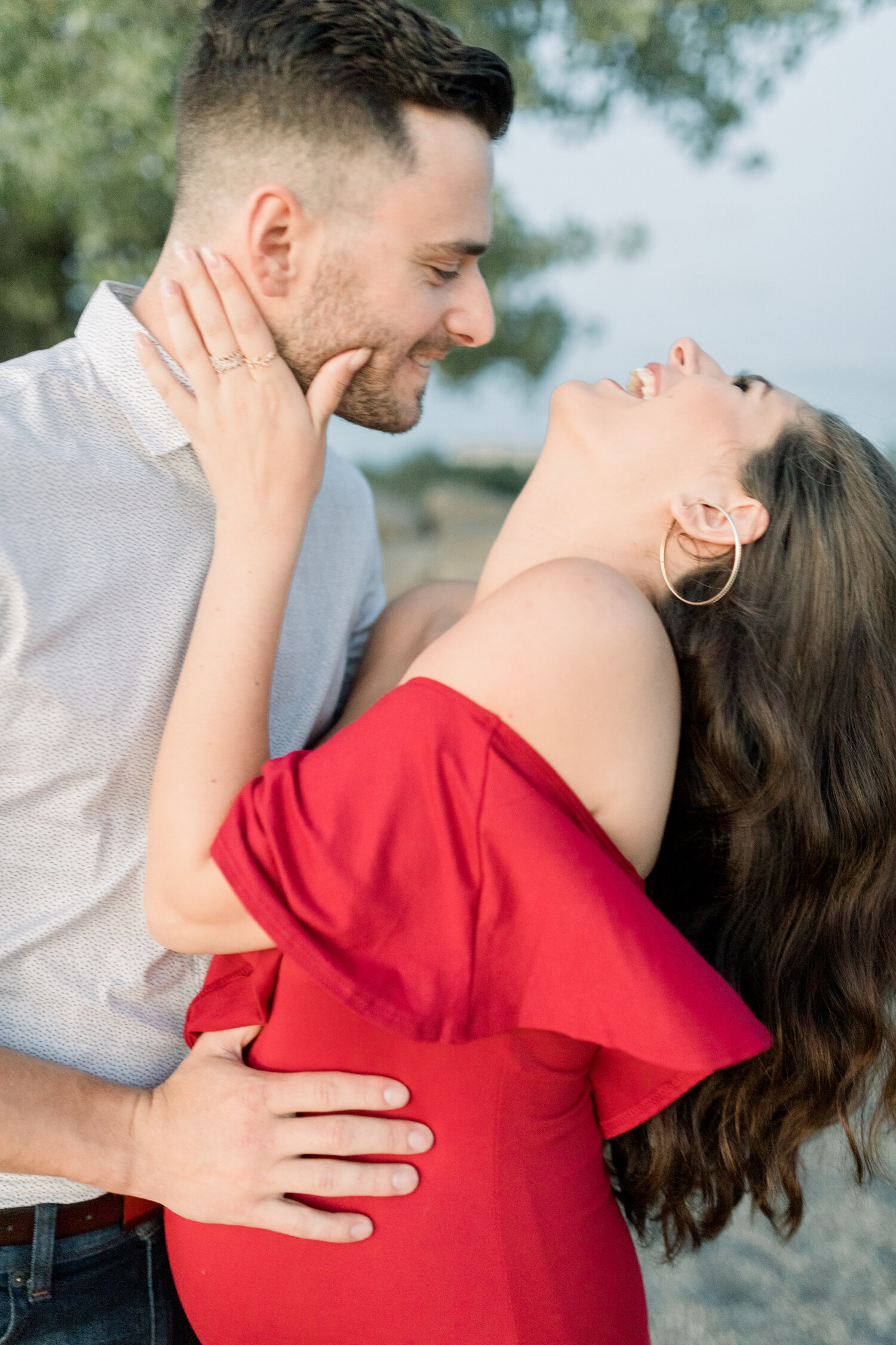  While embracing along the beach, Chelsea Mason Photography captures this expectant couple leaning their heads back and laugh during their maternity session in Ontario, Canada. off the shoulder red fitted maternity dress, woman leaning head back to l