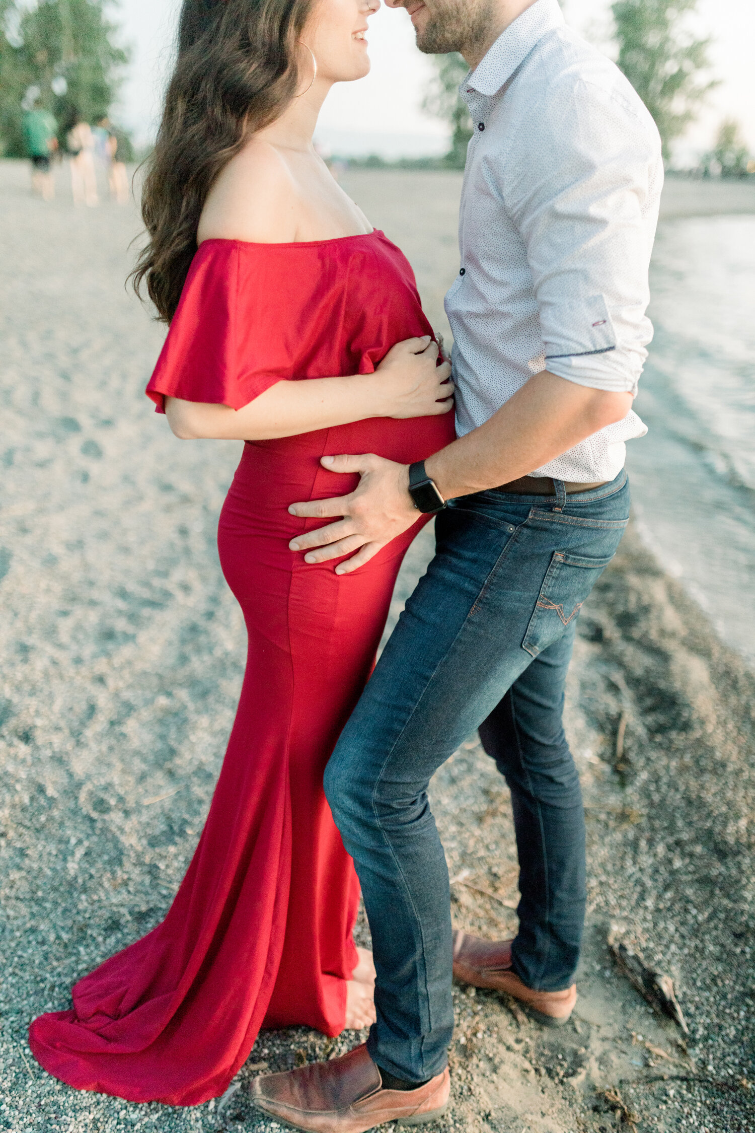  While standing along the shoreline of Brittania Beach in Ontario, Canada, Chelsea Mason Photography captures this expectant couple embrace one another. brittania beach, ontario photographer, maternity beach photos, red off the shoulder maternity dre