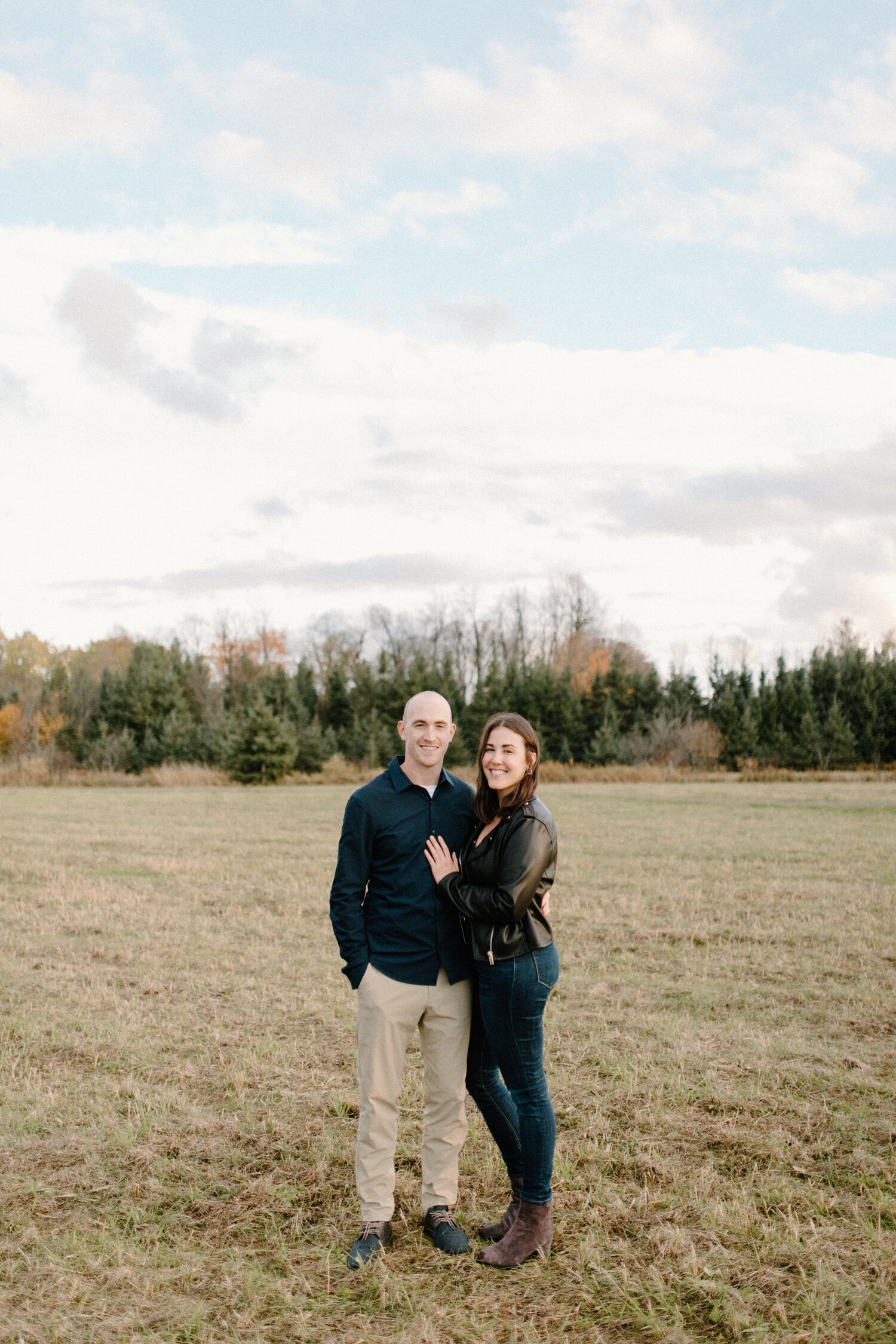  Posed in an open grass field in Ontario, Canada, Chelsea Mason Photography captures this fiance placing her hand on her soon-to-be groom’s chest and smiling. couple posing together womens leather black jacket mens khaki pants with black button up sh