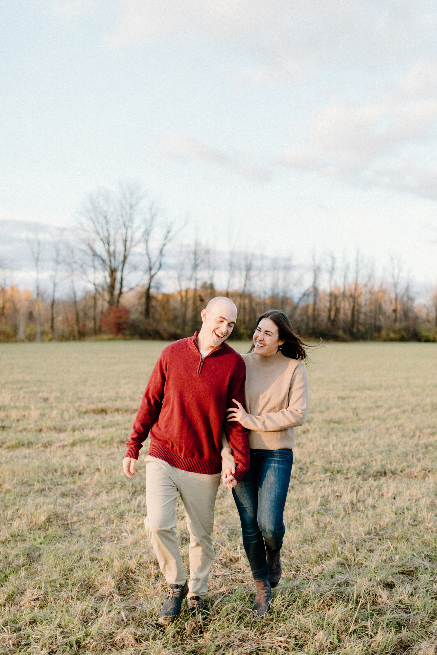  During their autumn engagement session in Ontario, Canada, Chelsea Mason Photography captures this couple linking arms and walking through an open field. autumn engagement session trees, mens red pullover with khaki pants, womens camel colored sweat