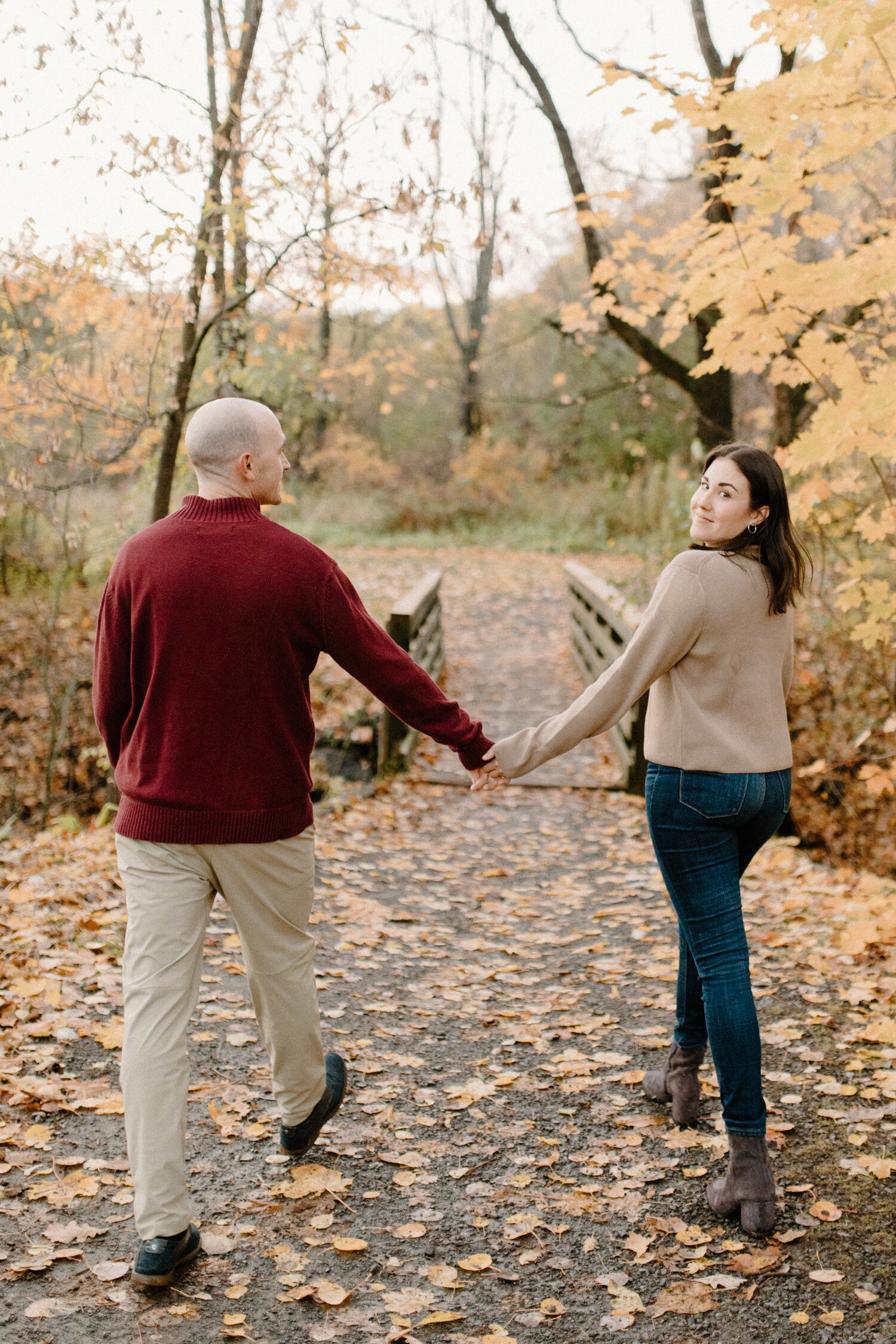  While walking on a wooden bridge in Ottawa Canada, hand-in-hand, this soon-to-be bride looks over her shoulder to smile for a photo for Chelsea Mason Photography. damp gravel ground with fallen autumn leaves, yellow autumn leaves, mens red pullover 