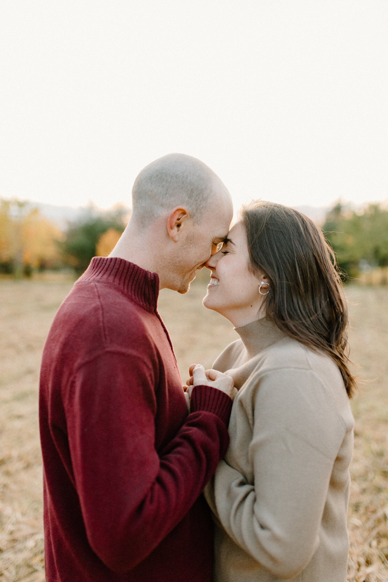  While posing in an open grass field in Ottawa, Canada, Chelsea Mason Photography captures this engaged couple holding hands tenderly and pressing their foreheads together. couple smiling and pressing foreheads together red and camel colored engageme