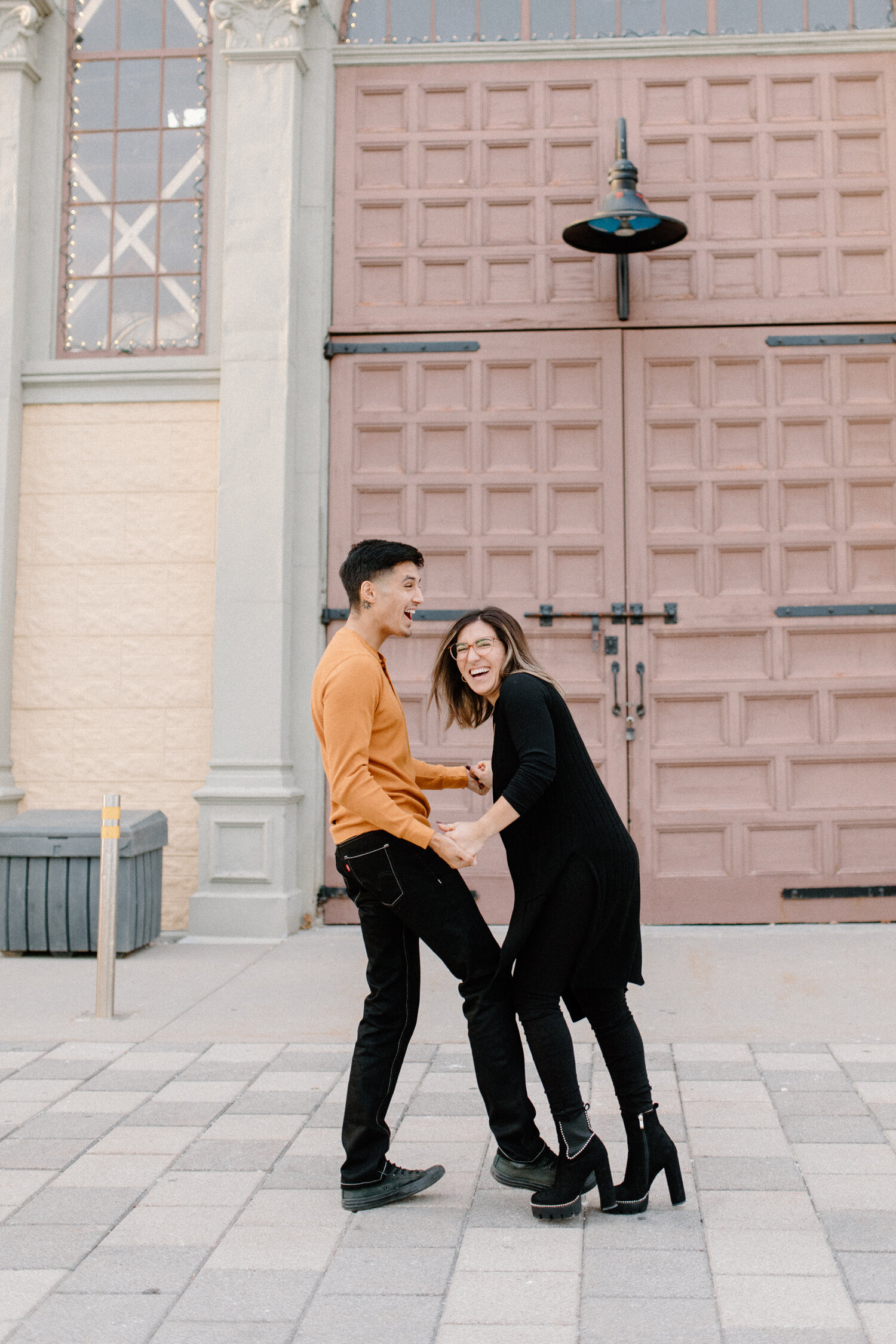  Chelsea Mason Photography captures this engaged couple holding hands outside the Ottawa, Canada Horticulture building. couple playfully holding hands womens all black outfit womens suede high heel booties mens orange and black outfit alternative cou