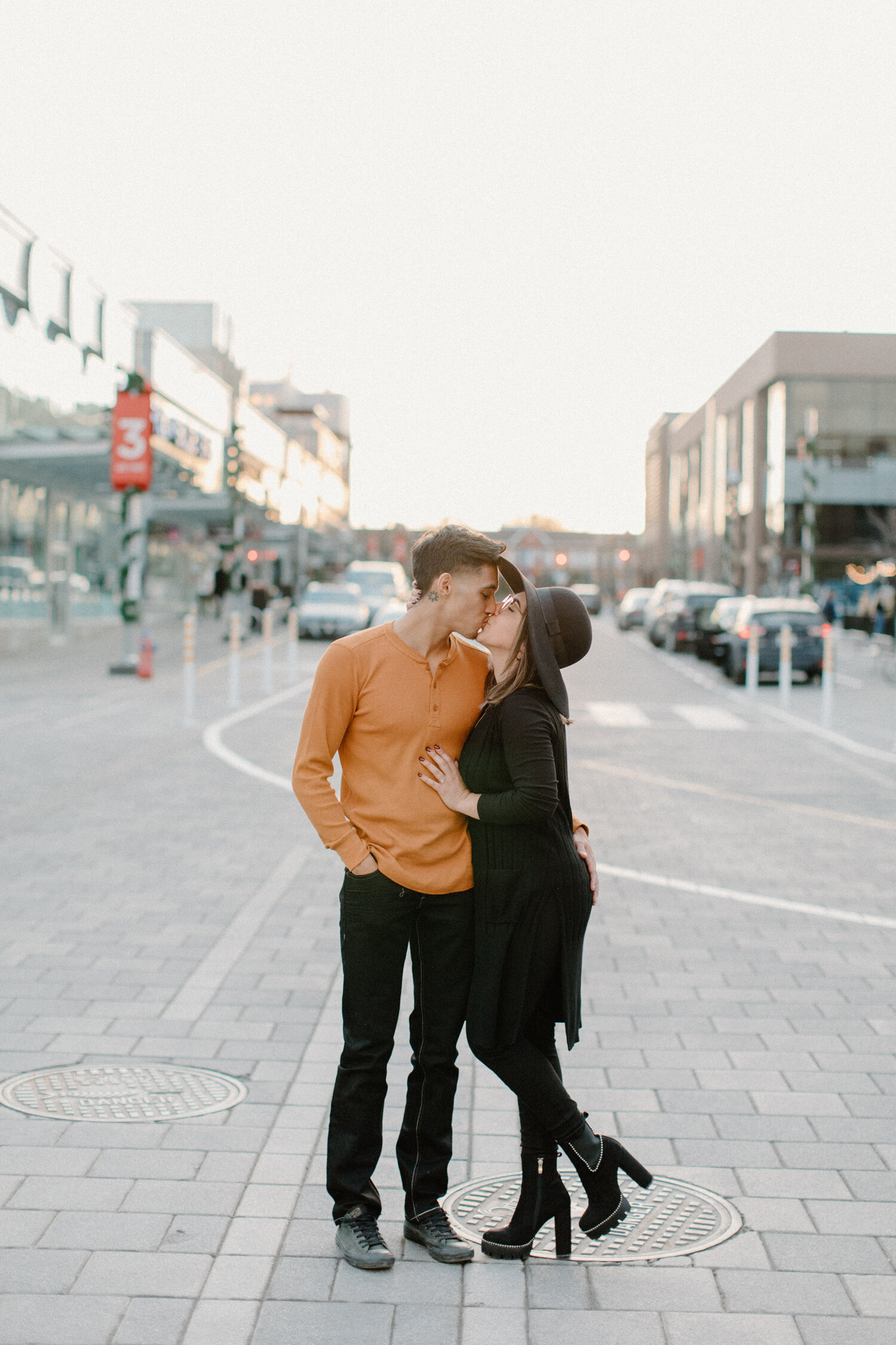  With urban shops blurred in the backdrop, Ottawa, Canada engagement photographer, Chelsea Mason Photography captures this couple holding each other close and kissing. womens thick heeled black sueded booties mens orange henley long sleeve shirt urba