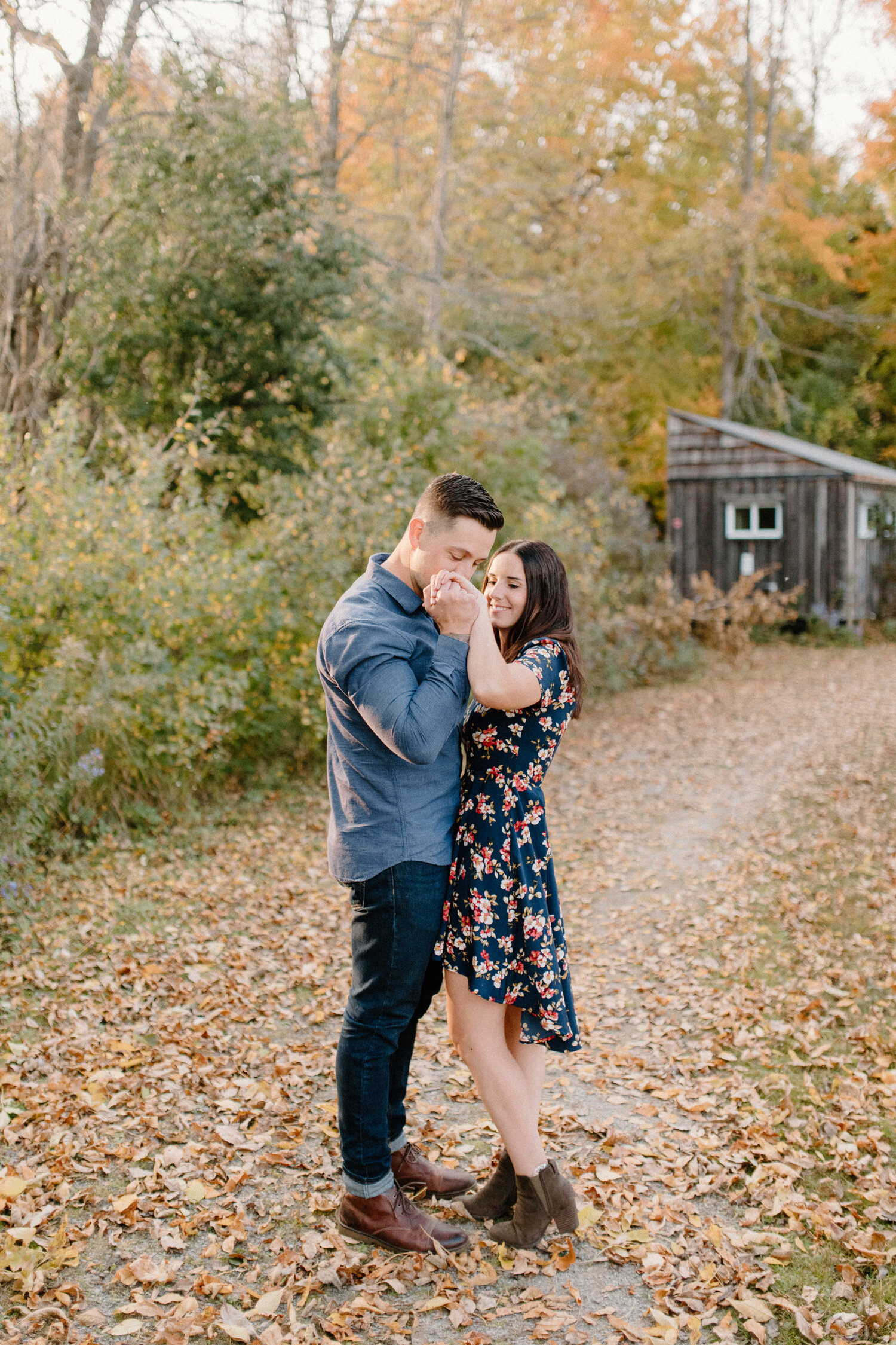  Outside a wooden cabin in the autumn woods of Brockville, Ontario, Chelsea Mason Photography captures this soon-to-be groom kissing his fiance’s hand. womens navy floral high low dress with brown suede booties mens denim outfit autumn fallen leaves 