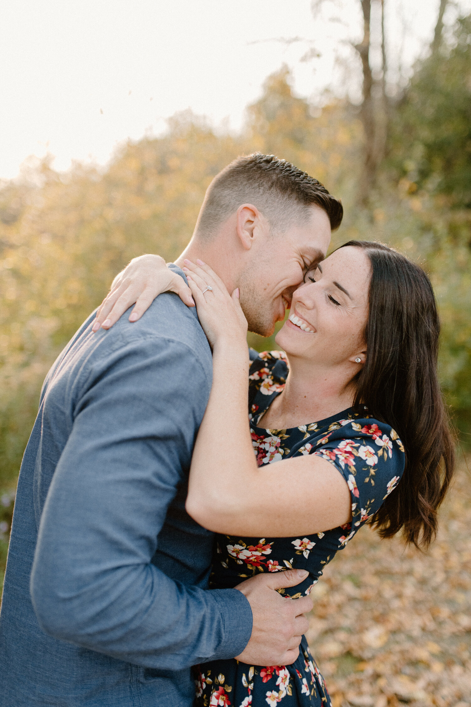  During their engagement session in Brockville, Ontario, this engaged couple wrapped their arms around one another while they smiled and laughed. man whispering into womans ear smiling playful engagement session engaged couple hugging and wrapping ar