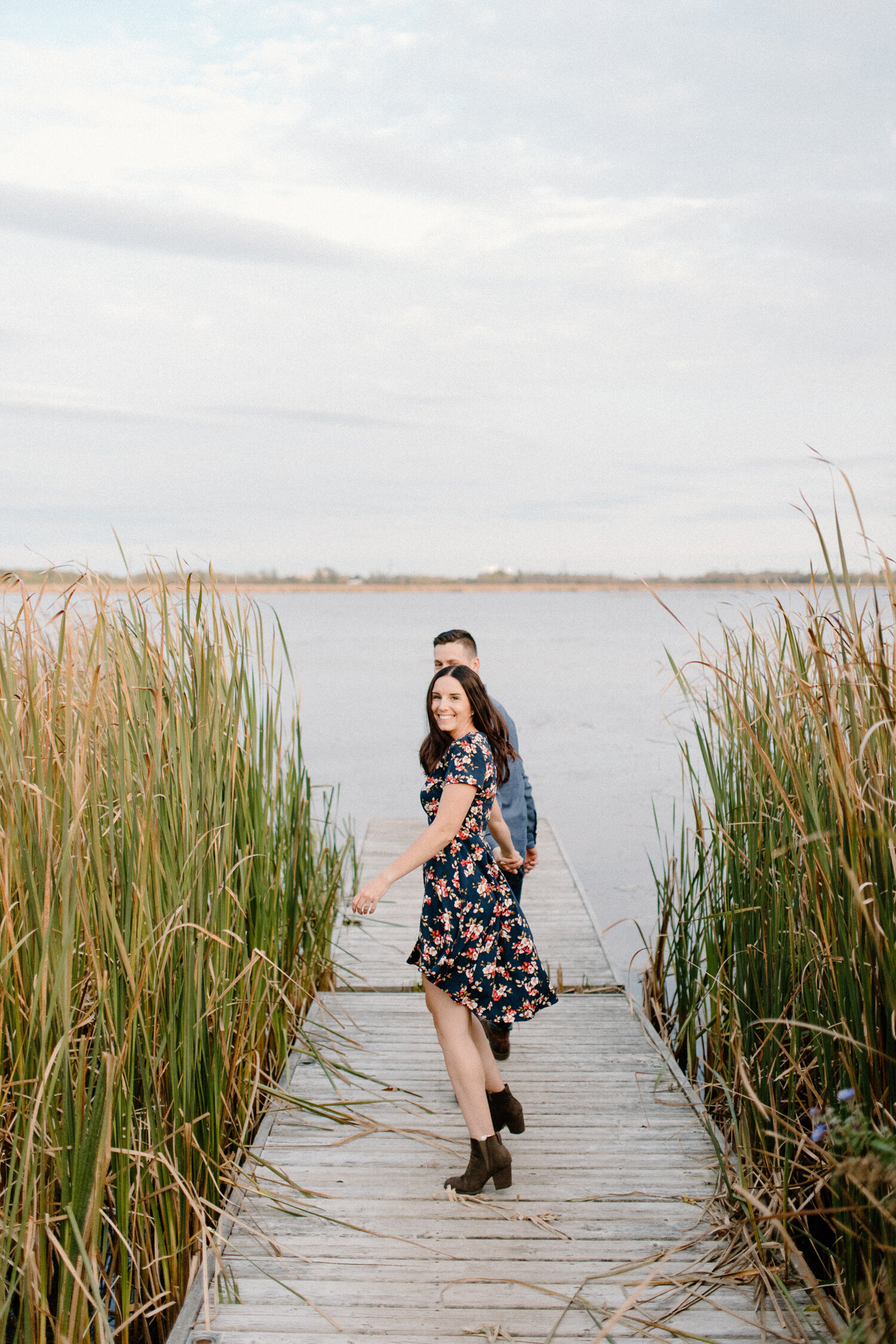  During this engagement session in Brockville, Ontario, Chelsea Mason Photography captures this engaged couple holding hands and walking onto a wooden lake dock. lake dock engagement session couple holding hands and smiling brockville ontario engagem