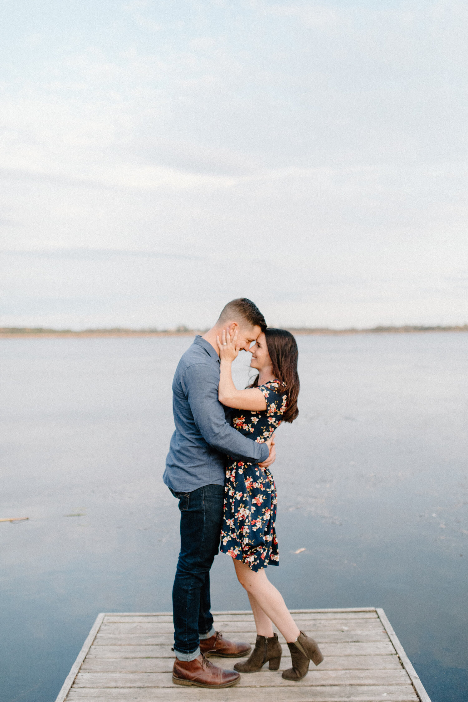  Chelsea Mason Photography captures this couple holding one another’s faces romantically on a pier in Brockville, Ontario. water pier engagement session wooden dock mens dark wash levi jeans with dark leather oiled shoes womens high low floral dress 