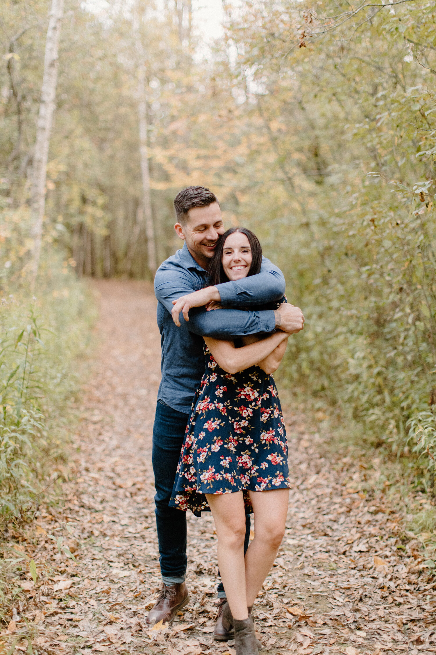  During this playful engagement photo session in Brockville, Ontario, Chelsea Mason Photography captures this couple laughing and wrapping their arms around one another on a hiking path. womens floral high low dress all denim mens outfit playful enga