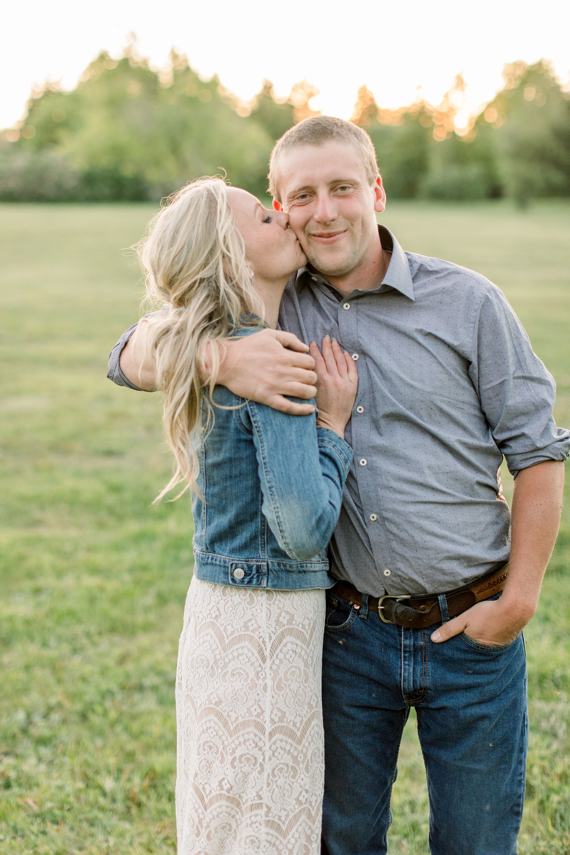  Posed in a grassy field in Ontario, Canada, Chelsea Mason Photography captures this engaged woman kissing her fiancé's cheek. engagement photographer Ottawa Ontario women's denim jacket with cream lace maxi dress southern styled engagement outfit  #