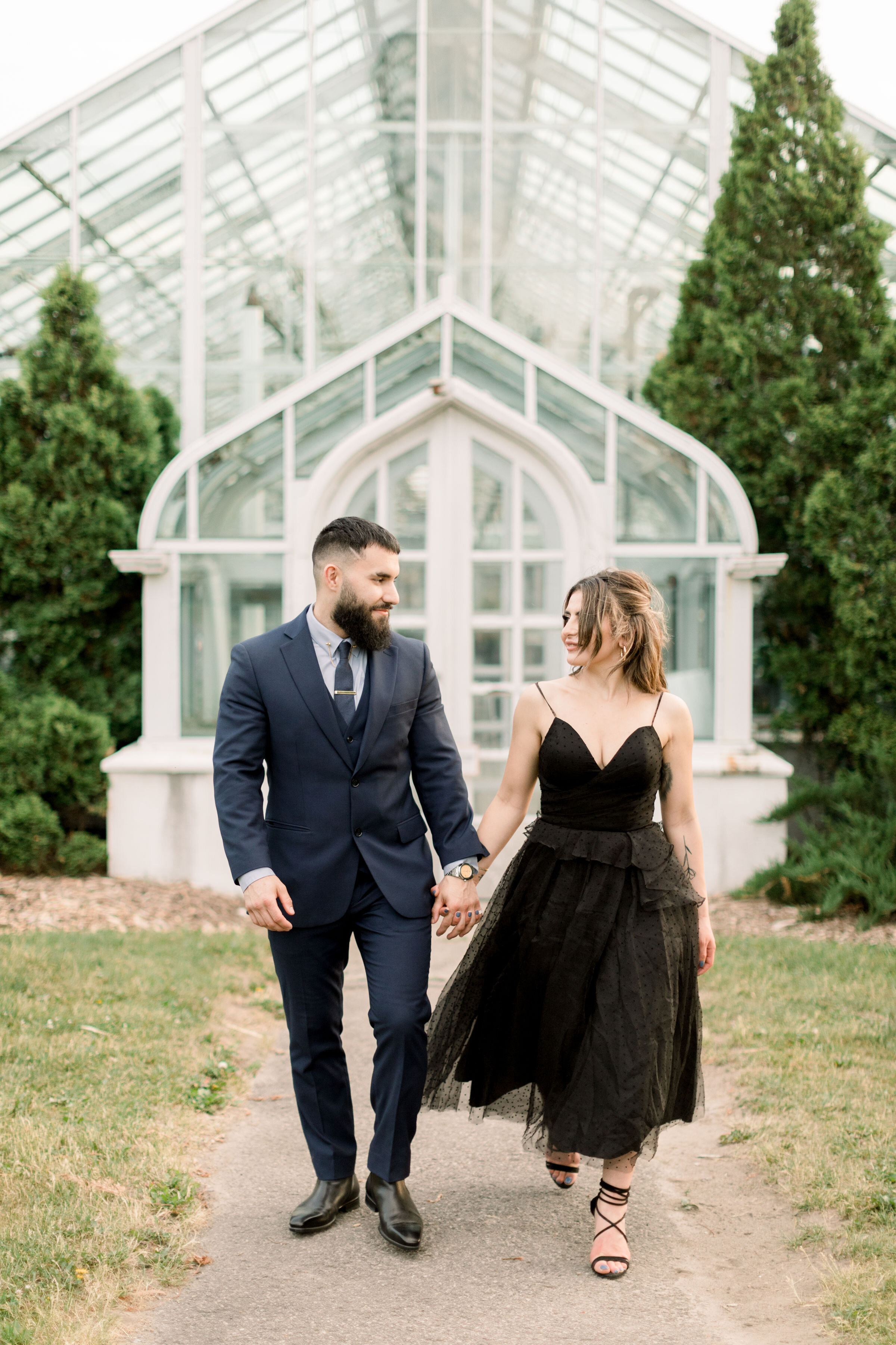  Chelsea Mason Photography captures this engaged couple walking hand-in-hand outside a beautiful white greenhouse in Ottawa, Ontario. greenhouse engagement session formal engagement outfit women's black spaghetti strapped tea length dress with scrapp