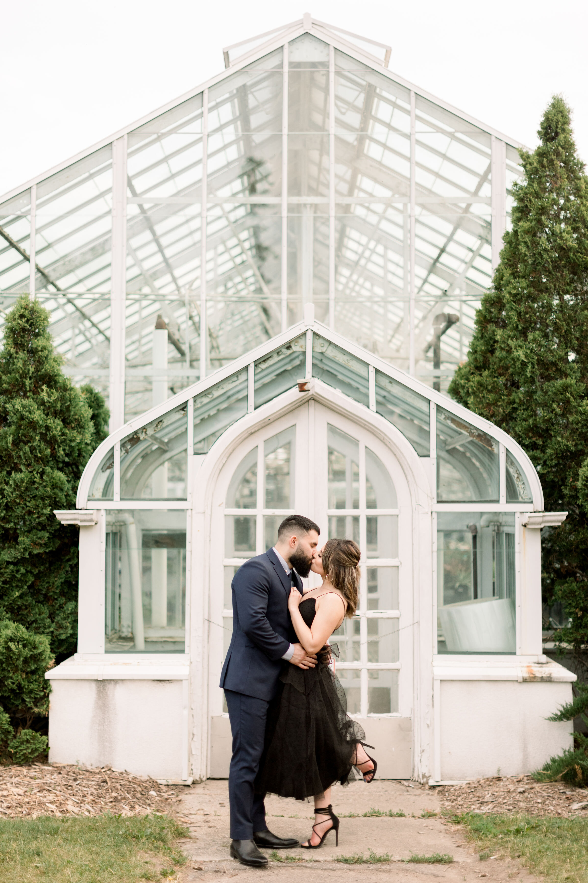  Chelsea Mason Photography captures this engaged couple romantically kissing with a beautiful white greenhouse in the backdrop in Ottawa, Ontario. greenhouse engagement session groom passionately kissing fiancé while she pops her foot #ChelseaMasonPh