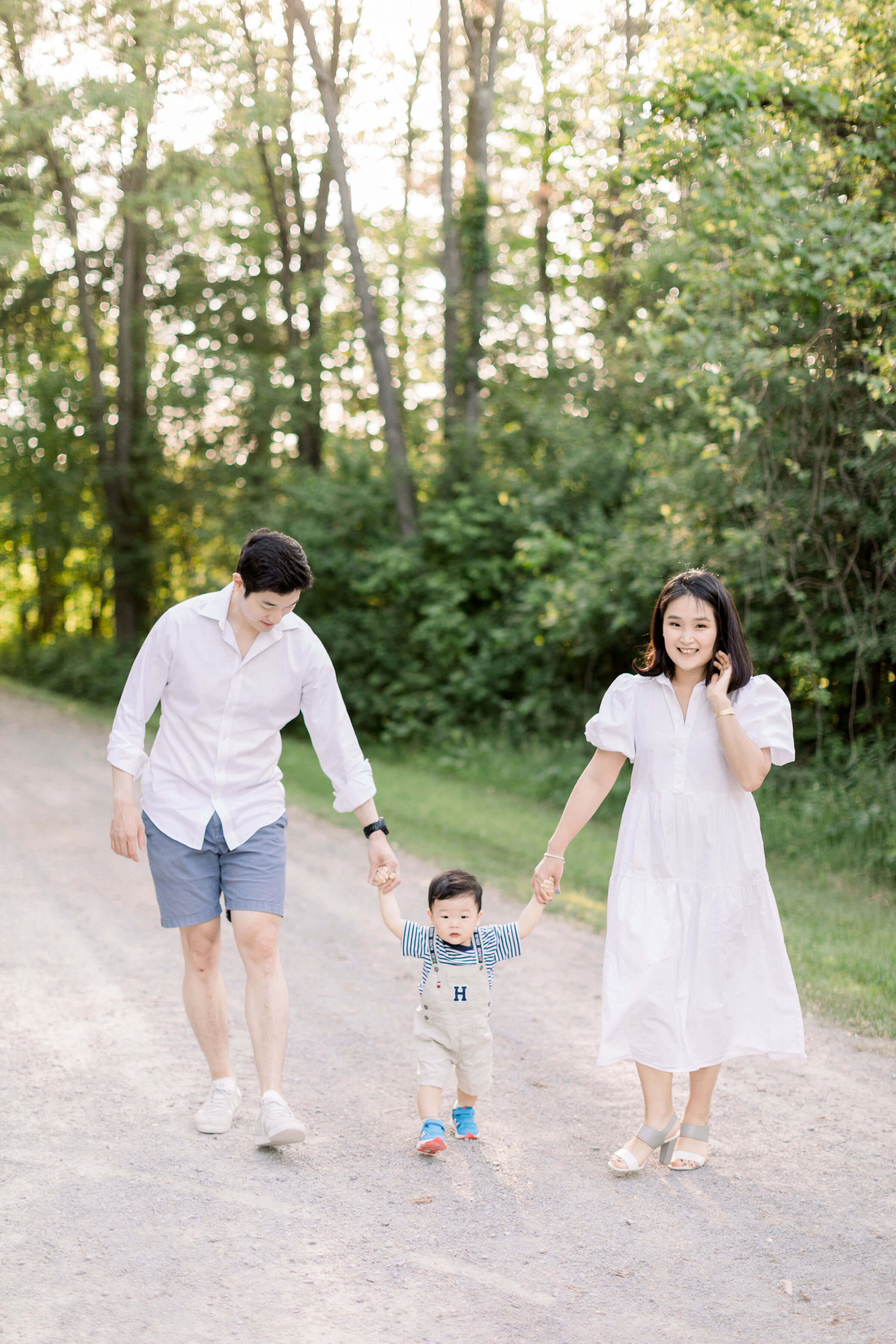  While walking down a wooded gravel pathway, Ottawa, Ontario family photographer, Chelsea Mason Photography captures these parents looking down at their toddler son as they hold his hands. family photo poses Ottawa family photographer #ChelseaMasonPh