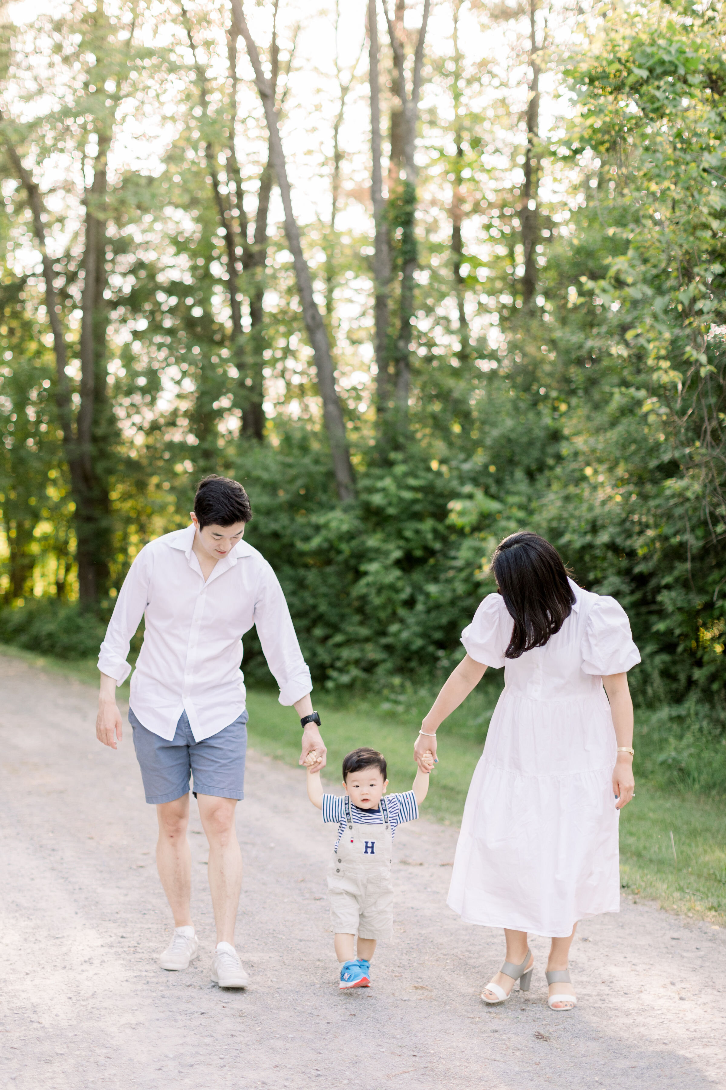  While walking down a wooded gravel pathway, Ottawa, Ontario family photographer, Chelsea Mason Photography captures these parents hold their toddler boy's hands. Ottawa family photographer white family photo outfit color toddler boy holding parents 